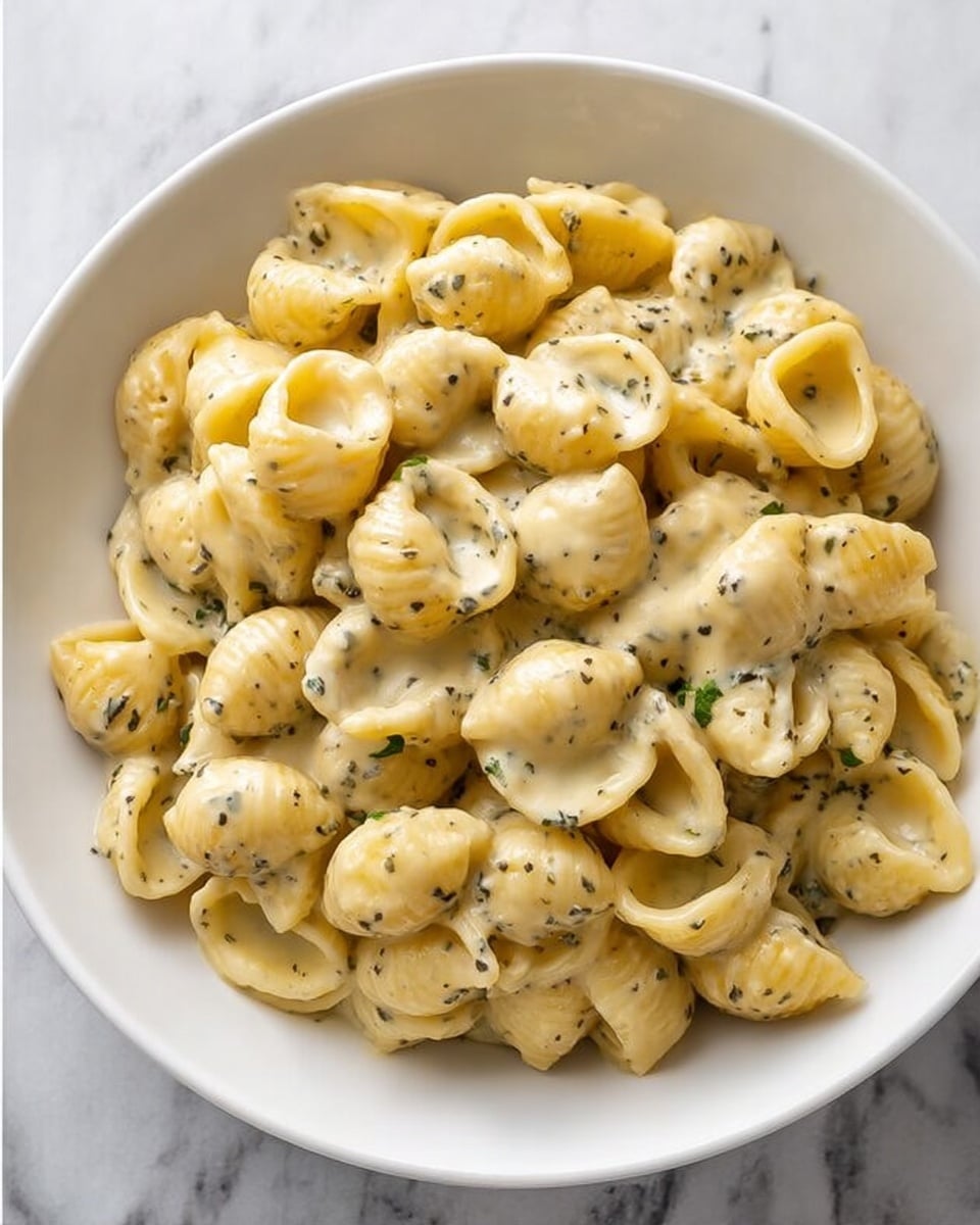 A white round bowl filled with a single layer of small shell-shaped pasta covered in a creamy, light yellow sauce with visible small dark green herb specks evenly spread throughout, sitting on a white marbled surface. The pasta shells appear soft and coated well by the sauce, giving a smooth and slightly shiny texture, making the dish look warm and comforting. Photo taken with an iphone --ar 4:5 --v 7