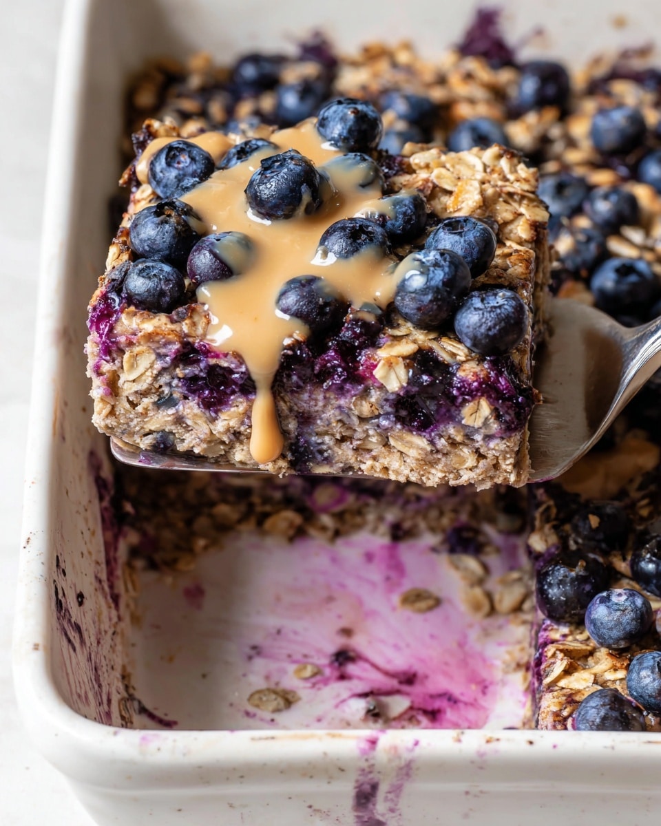 The image shows a square piece of baked oatmeal in a white baking dish, lifted by a silver spatula. The oatmeal has two main layers: a dense, purple-blue bottom layer filled with cooked blueberries, and a top layer covered with oats and fresh whole blueberries. Light brown sauce is drizzled unevenly over the top, adding a smooth texture. The white baking dish has some spots of purple blueberry juice and light stains around the edges, all set against a white marbled surface. photo taken with an iphone --ar 4:5 --v 7