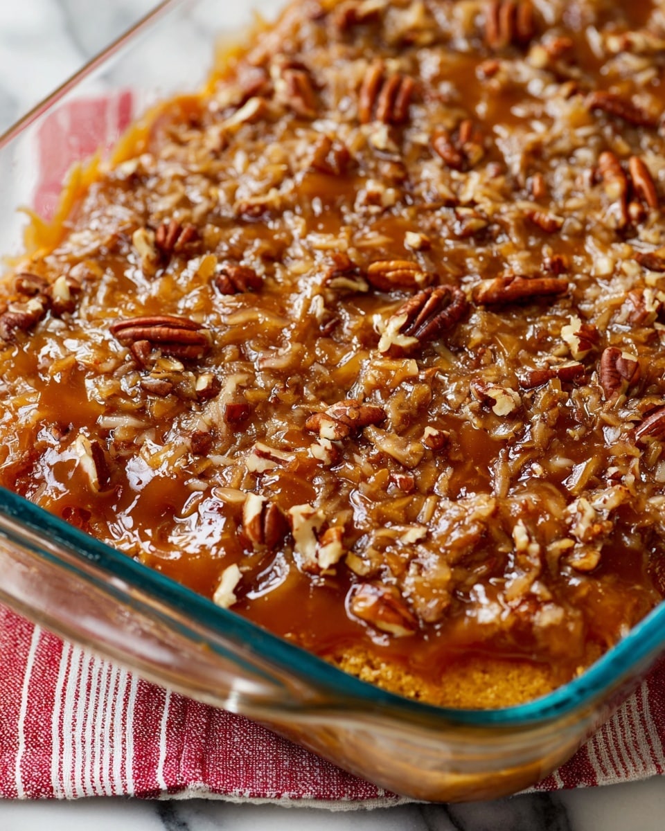 The image shows a close-up of a baked dessert in a clear glass dish, with one visible layer of golden-brown cake covered in a thick, glossy caramel-like sauce mixed with shredded coconut and chopped pecans. The sauce covers the entire surface, making it shiny and textured, with small pieces of nuts and coconut scattered evenly on top. The dish is placed on a white marbled surface with a red and white striped cloth partially visible underneath. Photo taken with an iphone --ar 4:5 --v 7