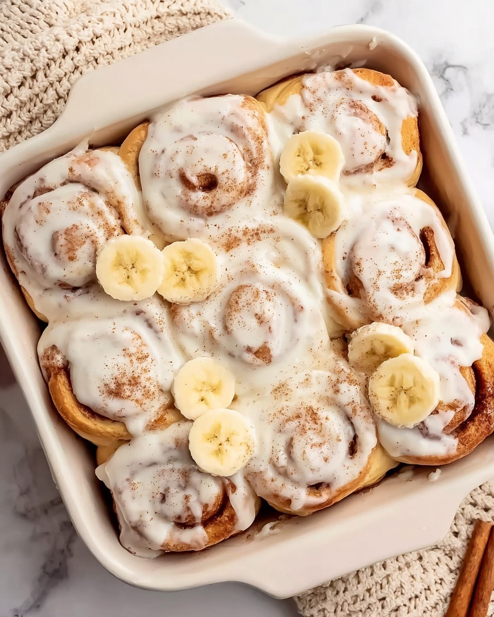 A white square baking dish holds ten cinnamon rolls covered in thick, creamy white icing that drips slightly over the edges of each roll. The rolls are arranged in three rows, with the bottom row having four rolls and the others three each, showing soft swirls of cinnamon brown peeking through the icing. On top, there are several round slices of fresh banana scattered unevenly, adding light yellow spots with a soft smooth texture. A light sprinkle of brown cinnamon powder dusts some areas of the icing. The dish sits on a white marbled surface, and a beige knitted cloth is partially visible to the side. photo taken with an iphone --ar 4:5 --v 7