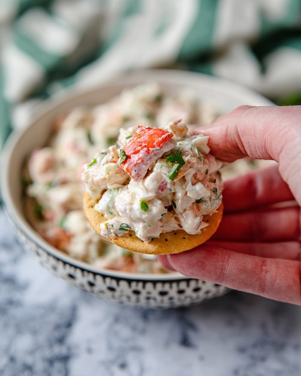 A woman's hand is holding a round, light brown cracker topped with a creamy seafood salad. The salad has white and light pink pieces mixed with small green herbs, and a bright red chunk of seafood is clearly visible on top. In the background, a white bowl with a black dotted rim is filled with more of the same creamy salad. A white marbled surface and a white and green striped cloth are softly blurred behind the bowl. photo taken with an iphone --ar 4:5 --v 7