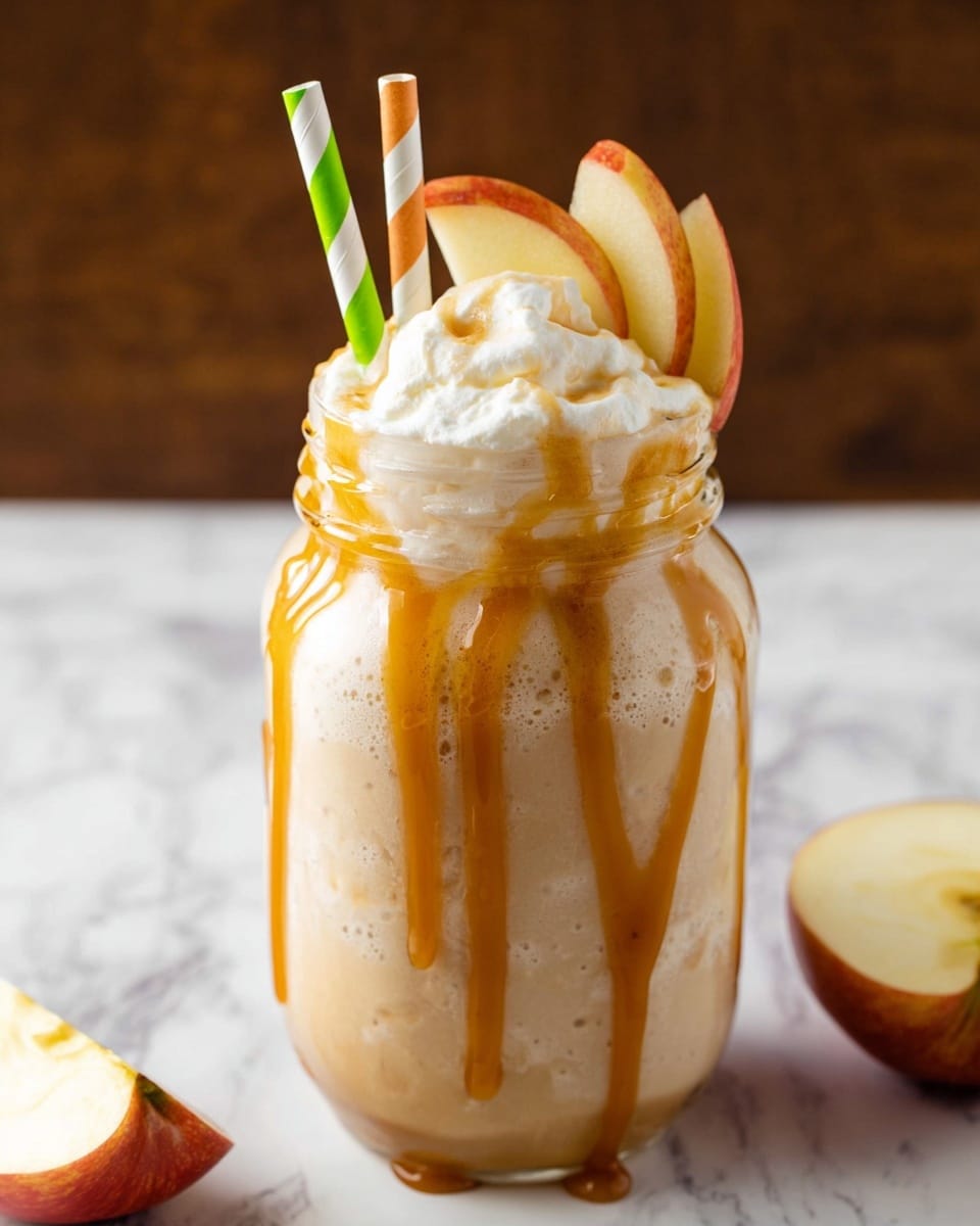 A clear glass jar filled with a light beige creamy drink, topped with a thick layer of whipped foam that slightly overflows the rim. Golden caramel sauce drips down the sides of the jar in thick streams. Two striped paper straws, one green and white and the other orange and white, are inserted into the drink. Thin slices of apple rest on top of the foam. The jar sits on a white marbled surface with an apple cut in half and a small apple slice placed nearby. Photo taken with an iphone --ar 4:5 --v 7