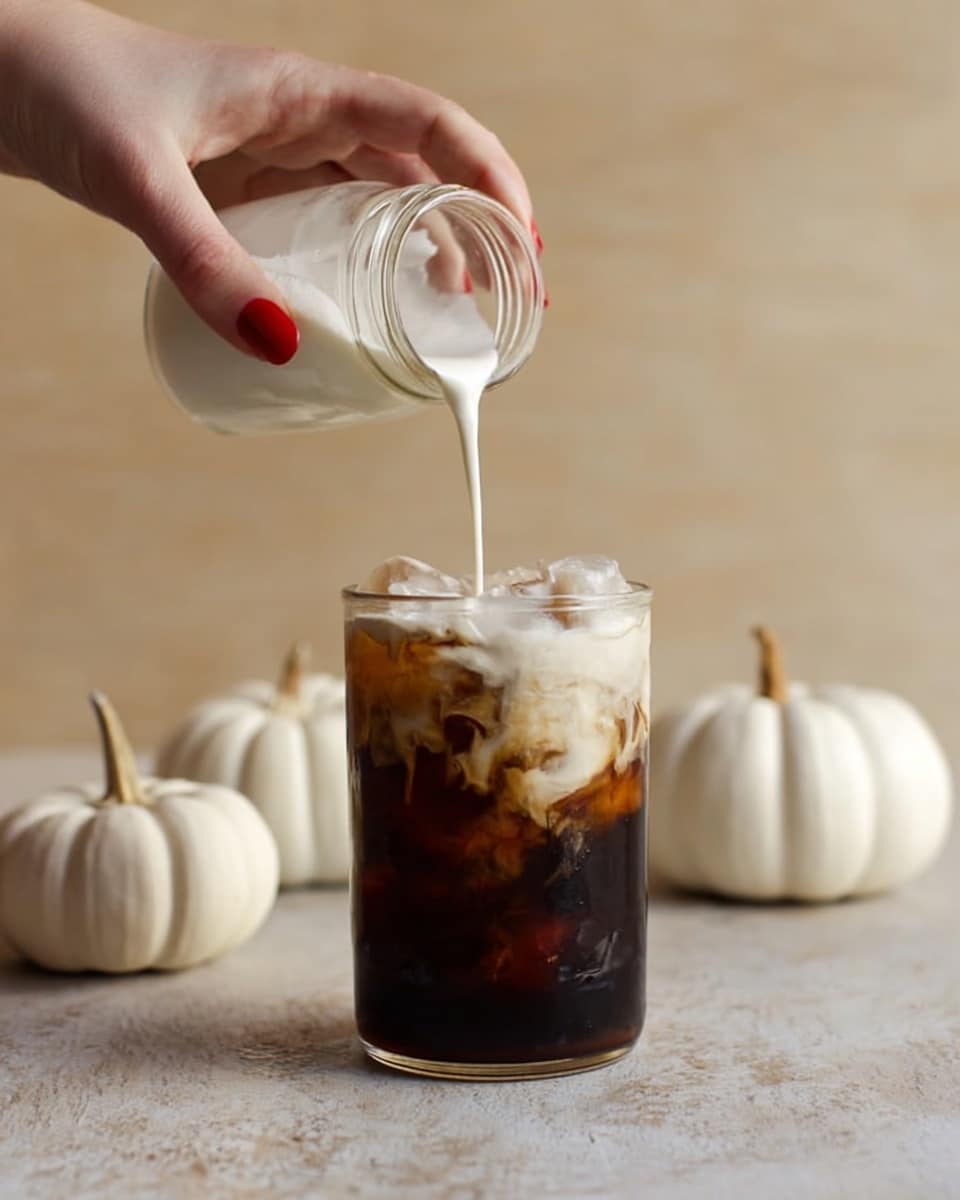 A woman's hand with red nail polish is pouring white creamy liquid from a small glass jar into a tall, clear glass filled with dark brown iced coffee and ice cubes; the creamy layer is thick and smooth, sitting on top of the dark iced coffee, showing a clear contrast between the two layers; the glass sits on a light tan textured surface with three small white pumpkins in the background; the overall tone is warm and cozy, with a soft light highlighting the textures. photo taken with an iphone --ar 4:5 --v 7