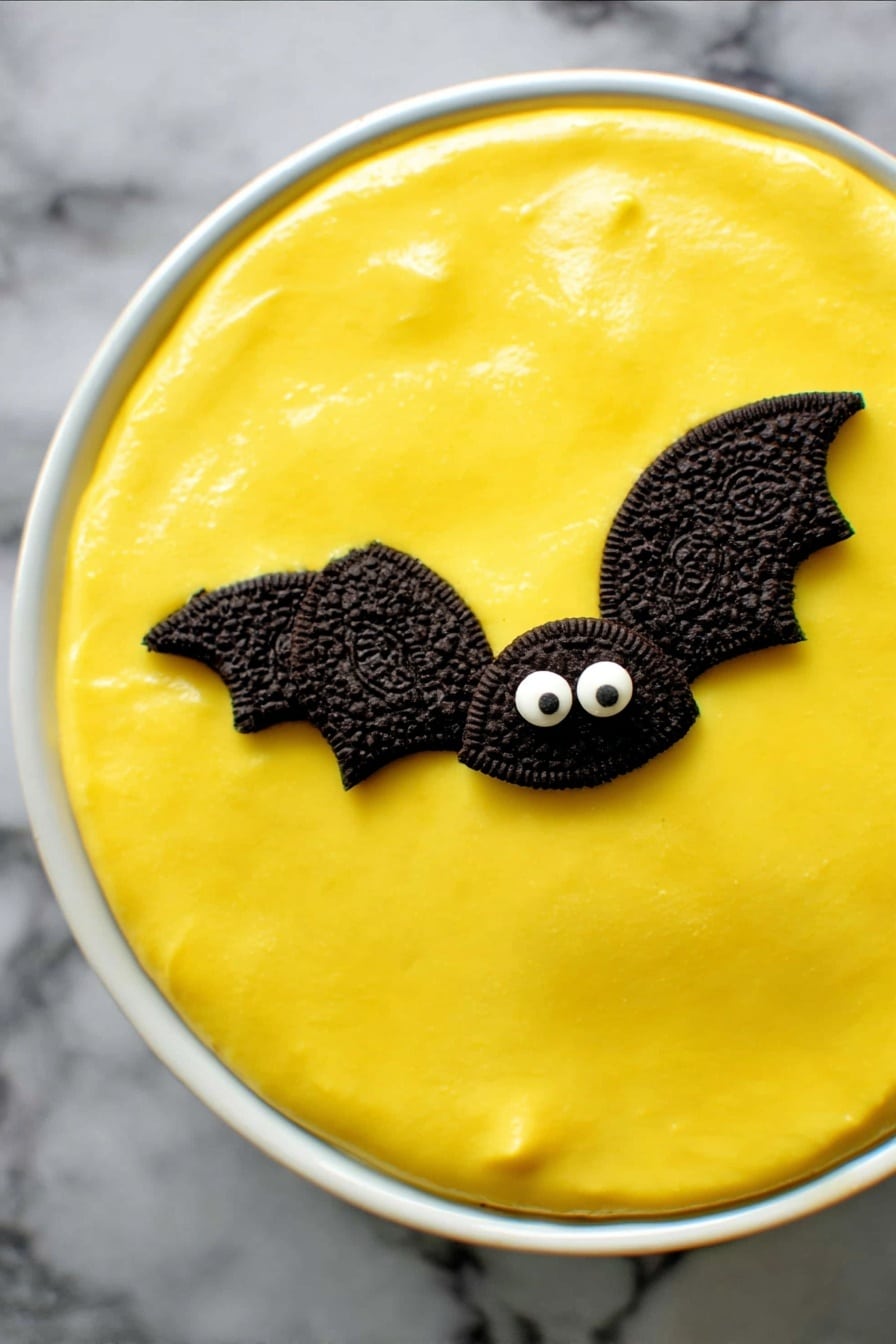 No Bake Boston Cream Halloween Pie, Halloween dessert, no bake pie recipe, spooky Halloween treat, easy Halloween dessert - A round dessert in a white ceramic dish has a smooth, bright yellow top layer that looks creamy and shiny. On this yellow surface are two small decorations shaped like bats, made with halves of dark chocolate sandwich cookies for wings and two small white candy eyes on a chocolate base forming the face. The dish sits on a dark cloth background. photo taken with an iphone --ar 2:3 --v 7