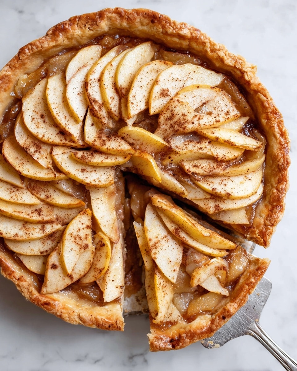 A round pie with a golden-brown crust that has a slightly rough texture and some cracks on the edges, filled with many thin, soft slices of pale yellow apple arranged in overlapping layers covering the whole surface. The apples are sprinkled with brown cinnamon powder, adding a warm color and texture contrast. Underneath the apple slices, there is a smooth, darker brown filling that looks like cooked fruit or syrup, giving the pie a glossy shine near the edges. One slice is cut and slightly pulled out with a silver pie server, showing the pie's layers clearly. The pie sits on a white marbled surface. photo taken with an iphone --ar 4:5 --v 7