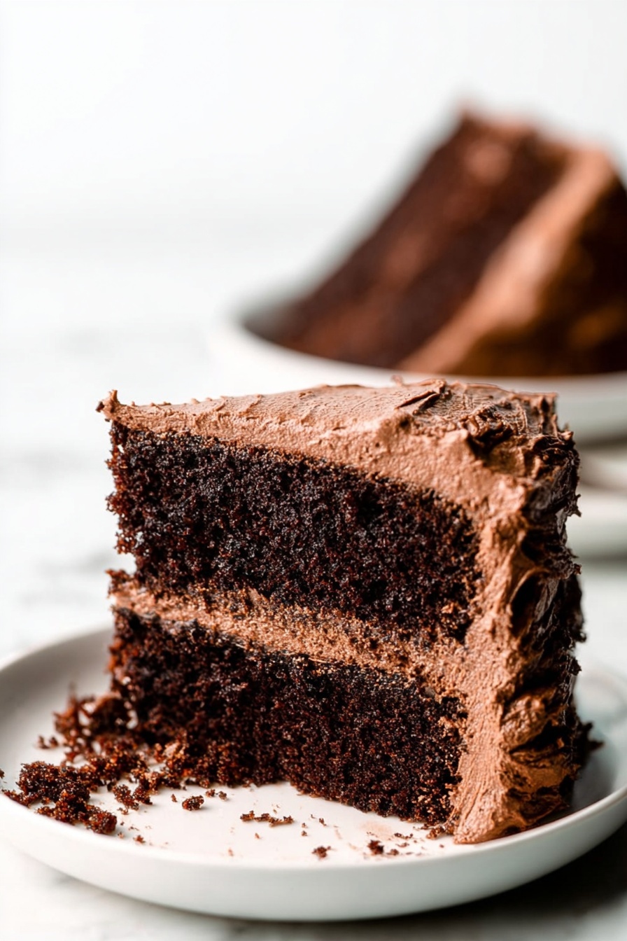 Gluten Free Vegan Chocolate Cake, vegan chocolate cake gluten free, dairy free chocolate cake recipe, easy gluten free vegan desserts, healthy vegan chocolate cake - A close-up view of a two-layer chocolate cake slice on a white plate, placed on a white marbled surface. The cake layers are dark brown and moist with a rough texture. Between the layers is a medium brown chocolate frosting that looks creamy and smooth. The outside of the cake slice is covered in the same chocolate frosting, which is thick and slightly swirled, with rough edges. Some frosting has spilled onto the plate. In the background, there is a blurred image of another chocolate cake slice on a white plate. Photo taken with an iphone --ar 2:3 --v 7