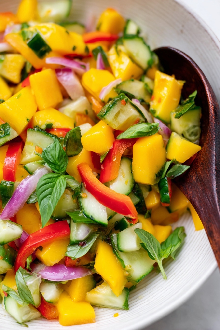 Mango Salad with Lemon-Honey Dressing, Mango Salad recipe, healthy fruit salad, easy summer salad, citrus mango salad - A close-up of a colorful salad in a white bowl with a white marbled background, showing bright yellow mango cubes, green cucumber pieces, red bell pepper chunks, thin slices of light purple onion, and fresh green basil leaves mixed together. A dark wooden spoon is resting inside the bowl, partly covered by the salad. The colors are vibrant and fresh, with the textures of the juicy mango, crunchy cucumber, and leafy greens clearly visible. Photo taken with an iphone --ar 2:3 --v 7