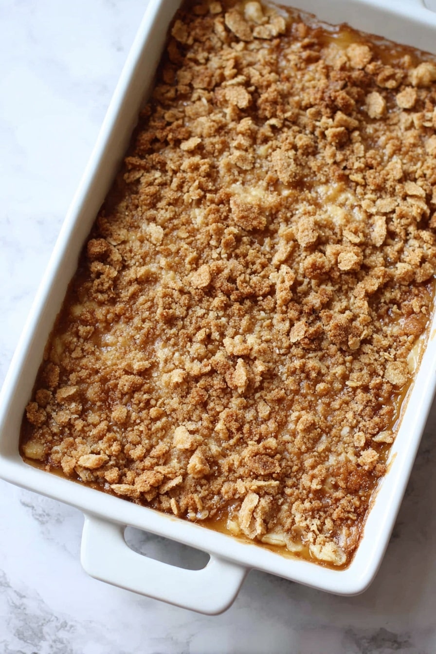 Pumpkin Dump Cake, easy pumpkin dessert, fall dessert recipes, quick dump cake, cozy pumpkin cake - The image shows a baked dessert in a white rectangular dish with two handles. The dessert has one main layer with a golden brown crumbly topping made of crushed pieces with a rough, uneven texture. The color is mixed shades of light to medium brown, giving a crunchy and toasted look. The dish is placed on a white marbled surface. Photo taken with an iphone --ar 2:3 --v 7