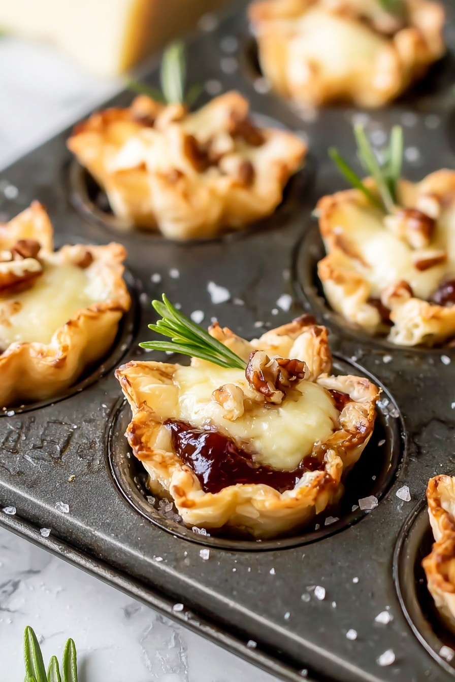 Cranberry Brie Bites with Puff Pastry, easy party appetizers, holiday appetizer recipes, flaky puff pastry snacks, cranberry brie finger foods - The image shows mini tartlets baked in a dark metal muffin tray, each tartlet having three main layers. The bottom layer is a golden-brown, flaky pastry shaped into small cups with slightly crispy edges. The middle layer is a glossy, dark red jam or fruit preserve that peeks out from beneath the top. The top layer is a melted, creamy pale yellow cheese, topped with small brown nut pieces and a fresh green rosemary sprig for garnish. The background is a white marbled texture, and some salt granules are scattered around the tray. photo taken with an iphone --ar 2:3 --v 7