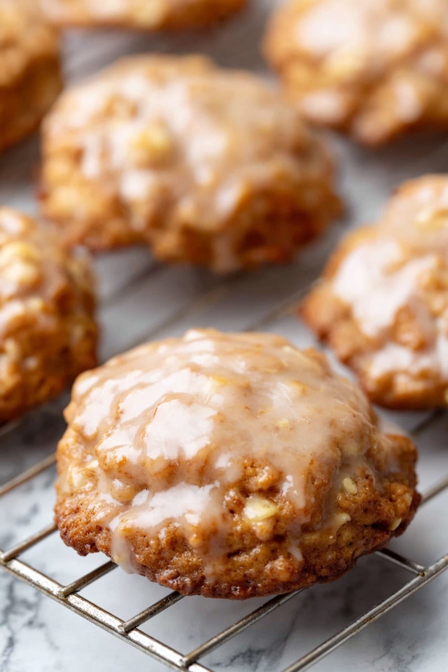 Crispy Apple Fritters with Sweet Vanilla Glaze, apple fritters, homemade apple fritters, cinnamon apple fritters, easy apple fritter recipe - The image shows several round cookies on a cooling rack. Each cookie has a shiny, light brown glaze on top with a slightly uneven texture. The cookies have a golden brown color, with a rough surface and visible small pieces inside, likely nuts or oats. The cooling rack underneath is metal and placed over a white marbled surface. The cookies look soft and have a slightly wavy edge. Photo taken with an iphone --ar 2:3 --v 7