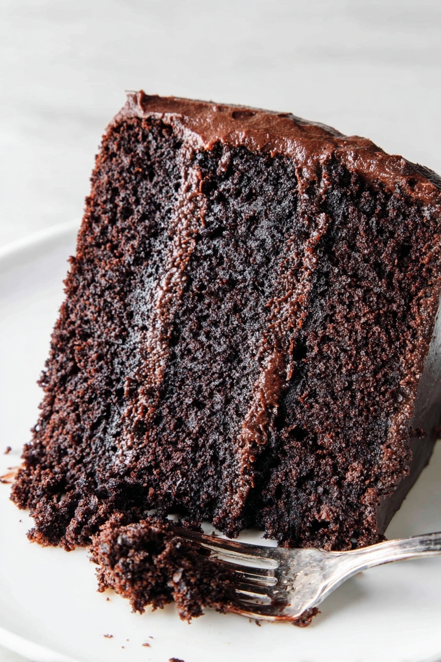The Most Amazing Chocolate Cake, chocolate cake recipe, moist chocolate cake, rich chocolate cake, homemade chocolate cake - The image shows a slice of chocolate cake on a white plate placed on a white marbled surface. The cake has two thick layers of moist, dark brown chocolate sponge with a rich, glossy chocolate frosting layer in the middle. The outside of the cake is covered with a smooth, shiny chocolate frosting. A silver fork with some chocolate frosting rests on the plate next to the slice. Photo taken with an iphone --ar 2:3 --v 7
