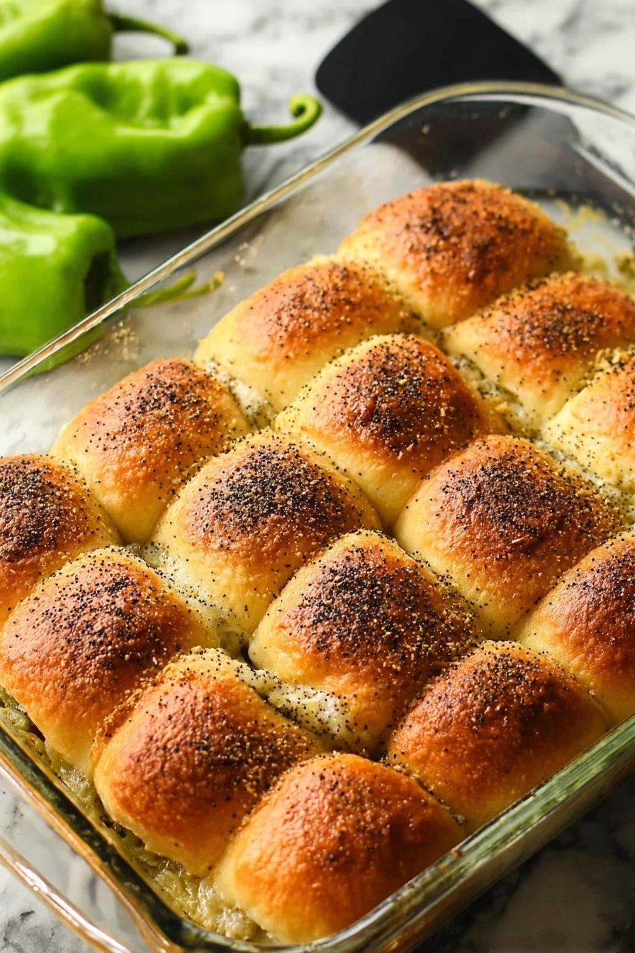 Santa Fe Turkey Sliders, Santa Fe Turkey Sliders recipe, spicy turkey sliders, Hawaiian roll sliders, easy party sliders - The image shows a clear glass baking dish filled with twelve golden brown sandwich rolls arranged in three rows of four. Each roll has a slightly crispy top with visible poppy seeds sprinkled over them, giving a textured look. The edges of the rolls touch each other, and a melted, creamy layer is seen bubbling around the sides beneath the rolls. In the background, there are some green fresh peppers resting on a white marbled surface. A black spatula is placed beside the dish. The photo is taken with an iphone --ar 2:3 --v 7