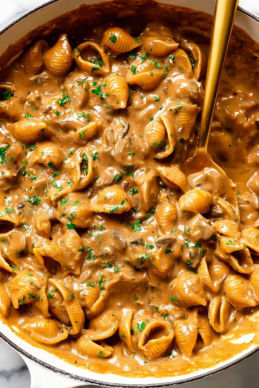 French Onion Pasta with Cheesy Sauce, French Onion Pasta, cheesy pasta recipe, caramelized onion pasta, easy comfort food pasta - A close-up view of a white pan filled with a rich, creamy mushroom sauce that has a warm brown color and a thick texture. The sauce contains many small mushroom slices that create a bumpy surface, covered evenly in the glossy, smooth sauce. Bright green parsley bits are scattered on top, adding a touch of color contrast. A shiny golden spoon rests inside the pan, partially submerged in the sauce, showing the thick consistency as it drags some sauce along. The background features a white marbled surface. photo taken with an iphone --ar 2:3 --v 7