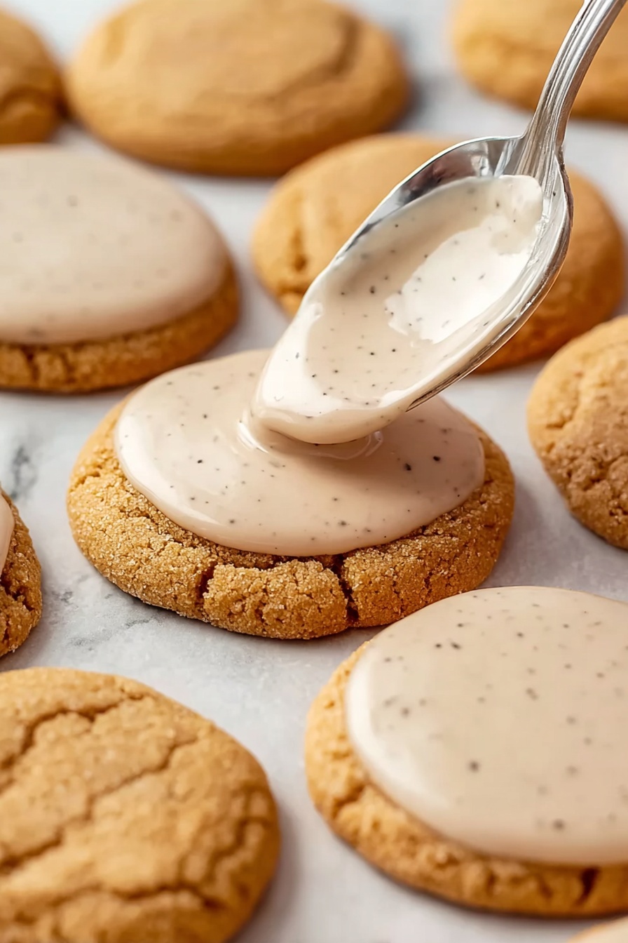 Brown Sugar Pop Tart Cookies, homemade pop tart cookies, cinnamon brown sugar cookies, soft buttery cookies, easy bake pop tart treats - Round light brown cookies with a soft, cracked surface are arranged on a white marbled background. A silver spoon is shown spreading a smooth, pale beige icing with small dark specks on the top surface of one cookie near the center. Several other cookies already have the same thin layer of icing evenly covering their tops while some remain plain. The cookies are placed closely together in rows and the icing appears glossy and creamy. Photo taken with an iphone --ar 2:3 --v 7