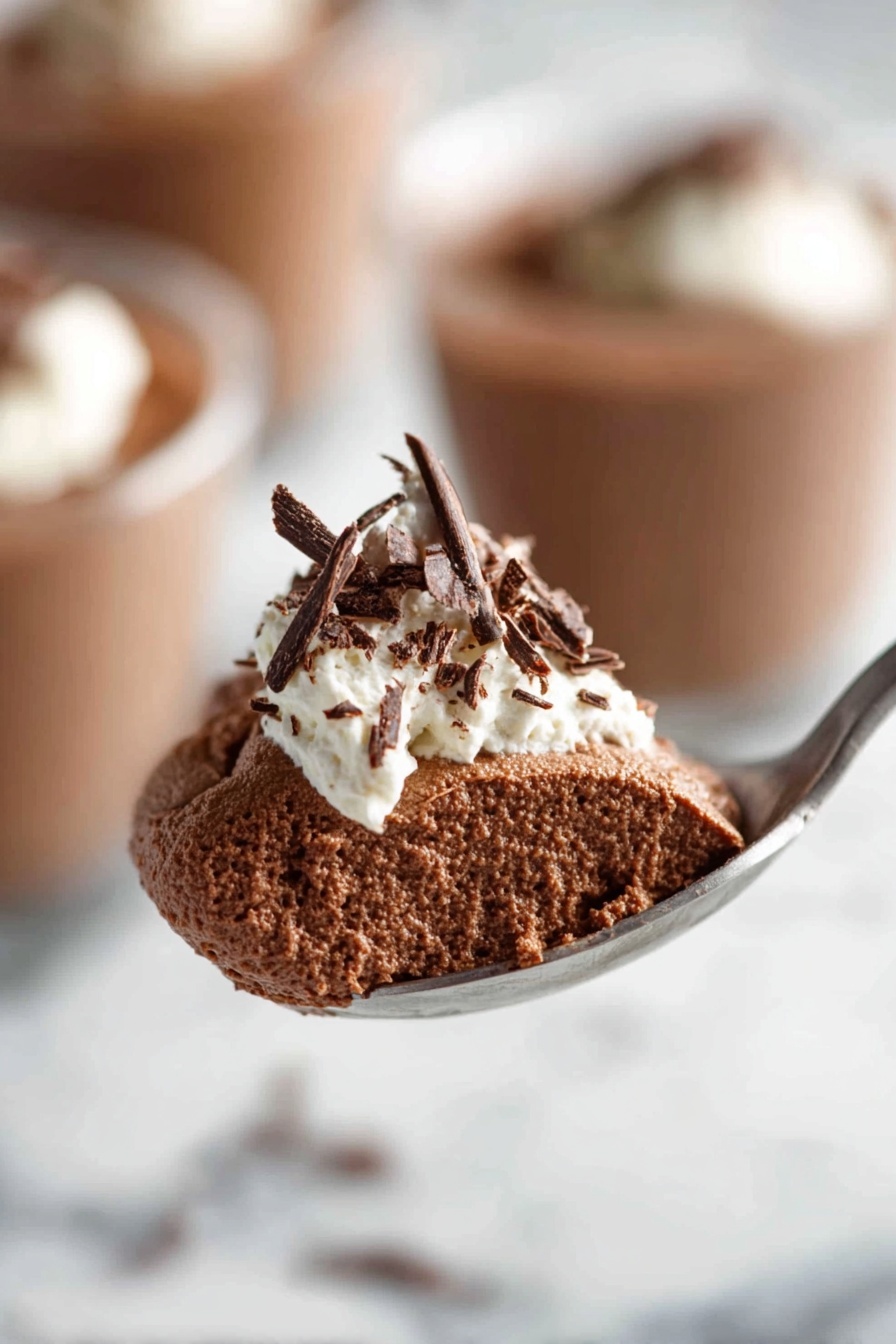 Decadent Chocolate Mousse, chocolate mousse recipe, rich chocolate dessert, easy chocolate mousse, creamy chocolate treat - The image shows a clear glass filled with a rich, smooth dark brown chocolate pudding. On top, there is a dollop of white whipped cream with delicate dark chocolate shavings scattered over it. The glass is placed on a white marbled surface, and in the background, there are blurred similar glasses filled with the same dessert. Photo taken with an iphone --ar 2:3 --v 7