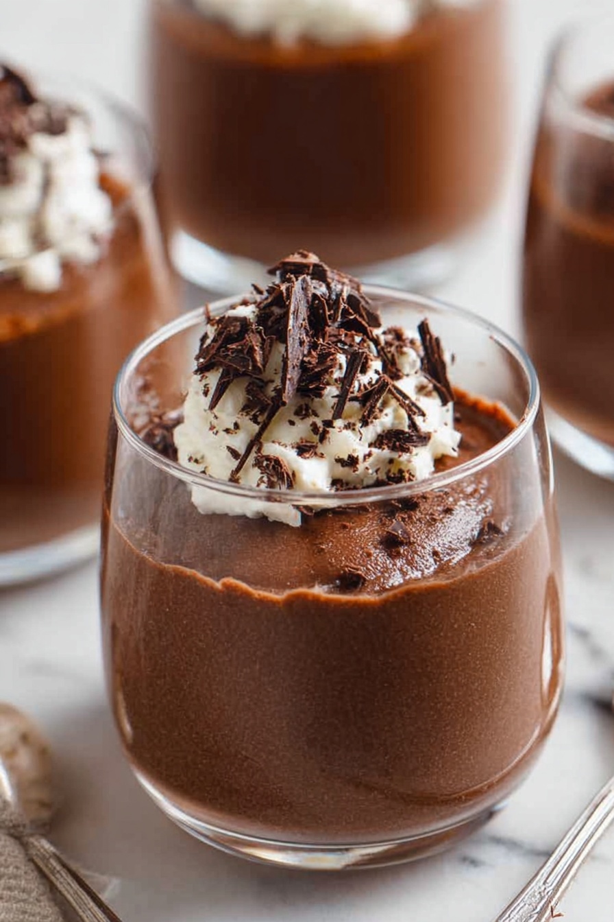 Decadent Chocolate Mousse, chocolate mousse recipe, rich chocolate dessert, easy chocolate mousse, creamy chocolate treat - A close-up of a spoon holding a scoop of light and airy chocolate mousse with a rough texture, topped with a small dollop of white whipped cream and thin, dark chocolate shavings scattered on top, with two blurry white bowls filled with the same mousse and cream in the background, all set on a white marbled surface. photo taken with an iphone --ar 2:3 --v 7