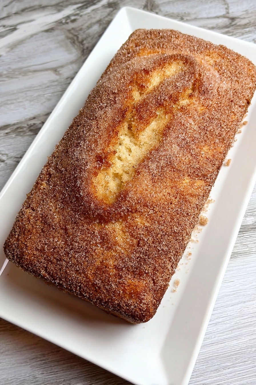 Cinnamon Donut Bread, cinnamon swirl bread, easy bread recipes, glazed bread loaf, homemade cinnamon bread - A rectangular cake with a golden-brown top layer covered evenly with a fine, grainy light brown sugar coating sits on a white rectangular plate. The cake's top surface shows a gently cracked texture with soft ridges and swirls, revealing a tender and moist interior below the sugar crust. The plate rests on a white marbled texture surface. photo taken with an iphone --ar 2:3 --v 7