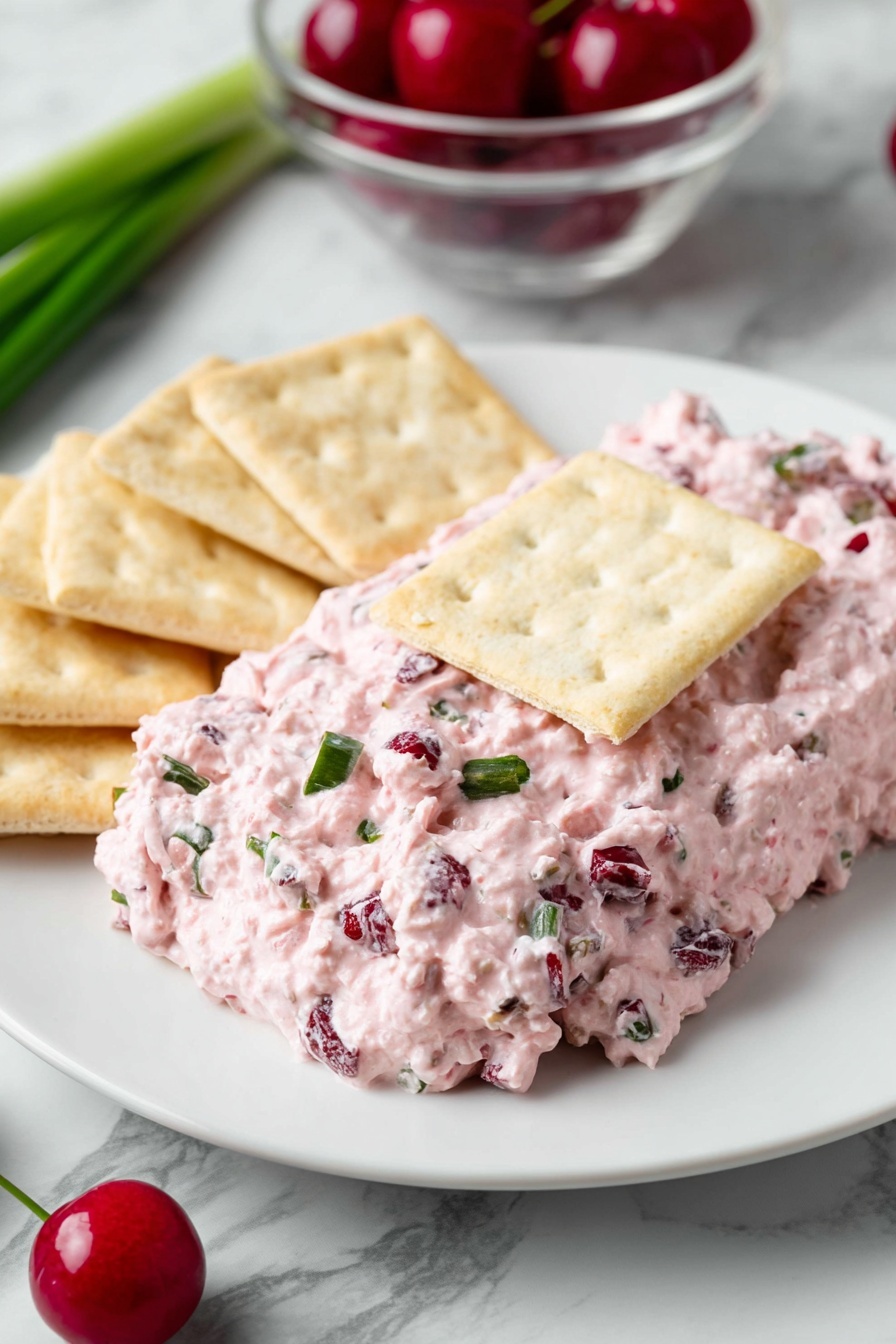 Cranberry Jalapeno Dip, Cranberry Jalapeno Dip flavor, vegan Cranberry Jalapeno Dip, easy Cranberry Jalapeno Dip, festive Cranberry Jalapeno Dip - On a white plate, there is a thick layer of pink creamy spread with small chunks of darker red and green mixed in. The spread is topped with a rectangular light beige cracker in the center. Around the spread, there are more light beige crackers placed on the white plate. The background shows a green onion on a white marbled surface and a clear bowl with red cherries. A green chili pepper is partially visible near the plate. photo taken with an iphone --ar 2:3 --v 7