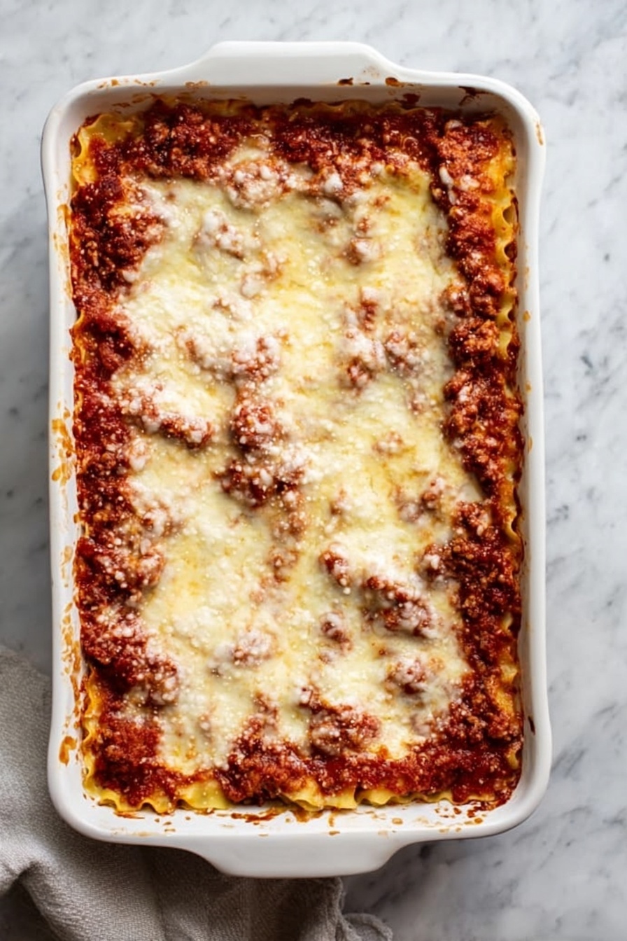 Vegan Lasagna Rolls, vegan lasagna, plant-based lasagna, dairy-free lasagna, vegan Italian dinner - A white rectangular ceramic baking dish filled with a baked lasagna, showing at least three layers: a bottom layer of pasta sheets, a middle layer of rich red tomato sauce mixed with browned ground meat, and a top layer covered evenly with melted, slightly browned cheese that looks soft and smooth. The edges of the dish have some browned sauce splashes. The dish sits on a surface with a white marbled texture, and a light-colored cloth is partially visible under the dish on the lower left side. Photo taken with an iphone --ar 2:3 --v 7