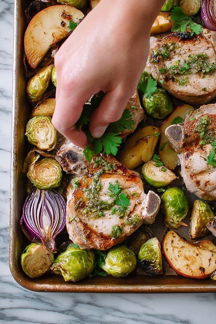 Sheet Pan Lemon Sage Pork Chops with Roasted Apples and Brussels Sprouts, Lemon Sage Pork Chops, Easy Sheet Pan Dinner, Roasted Apple Brussels Sprouts, One-Pan Pork Chop Recipe - A close-up view of a metal tray filled with cooked pork chops and roasted vegetables. There are three pork chops, each light brown with grill marks and green herb sauce on top, scattered across the tray. Around the pork chops, there are halved Brussels sprouts with a charred, darker green color, quartered pieces of red onions with purple layers, and golden brown apple slices. A woman's hand is gently placing fresh green parsley leaves over the pork chops. The tray sits on a white marbled surface. photo taken with an iphone --ar 2:3 --v 7