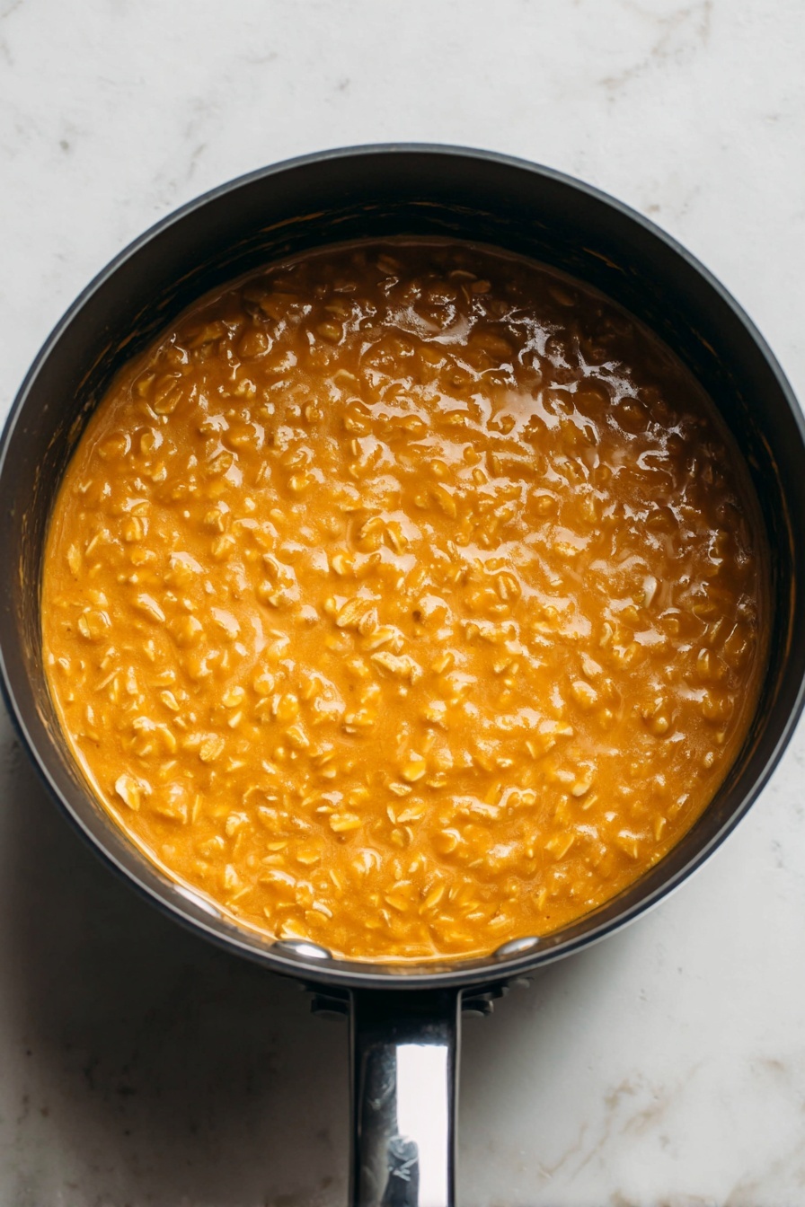 Pumpkin Oatmeal with Spiced Toppings, cozy breakfast recipes, healthy pumpkin oatmeal, seasonal fall breakfast, easy pumpkin oatmeal - This image shows a close-up top view of a round black saucepan filled with a thick, creamy orange mixture with visible small oats throughout. The oats give the surface a bumpy texture, and the mixture looks smooth and warm. The saucepan is placed on a white marbled surface, and the shiny black handle is visible at the bottom right of the image. The lighting highlights the glossy shine of the mixture, making it look fresh and appetizing. photo taken with an iphone --ar 2:3 --v 7