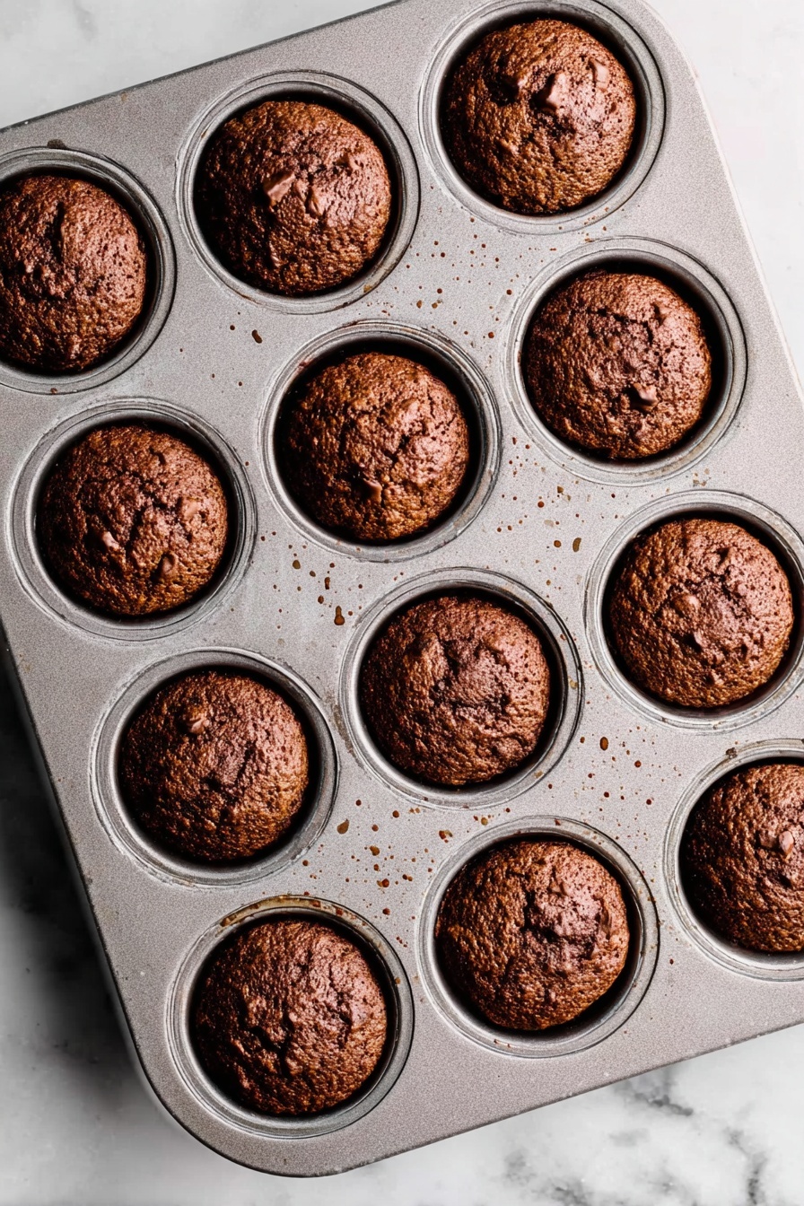 Sticky Toffee Pudding with Warm Toffee Sauce, how to make sticky toffee pudding, easy sticky toffee pudding recipe, best toffee pudding dessert, moist sticky toffee pudding - A metal muffin tray with 12 evenly spaced round muffins baked inside, each muffin a deep brown color with a rough, slightly cracked top texture showing some small chunks within. The muffins fit neatly in each circular slot of the tray, which shows small spots of light brown residue from baking. The tray is placed on a white marbled surface with soft natural lighting highlighting the muffins' golden and dark brown hues. photo taken with an iphone --ar 2:3 --v 7