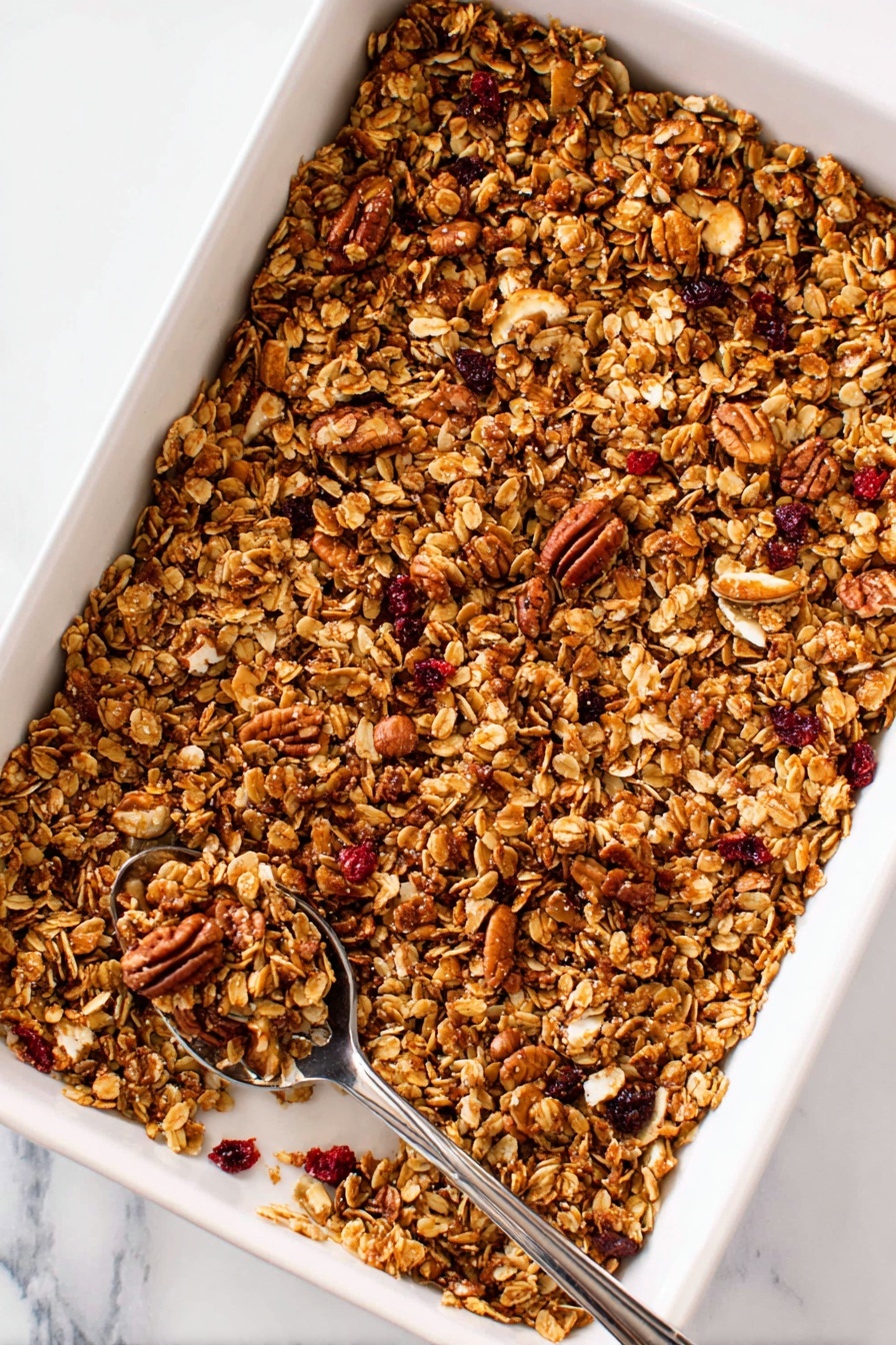 Gingerbread Granola, gingerbread granola bars, cozy breakfast granola, seasonal homemade granola, spiced crunchy granola - A deep white rectangular baking tray filled with a golden brown granola mix that includes oats, nuts like pecans, and small red dried fruit pieces scattered evenly throughout. The granola layer looks crunchy with a mix of small and larger clusters, and a silver spoon rests on the bottom left corner inside the tray, holding some granola pieces. The background is a white marbled surface. Photo taken with an iphone --ar 2:3 --v 7
