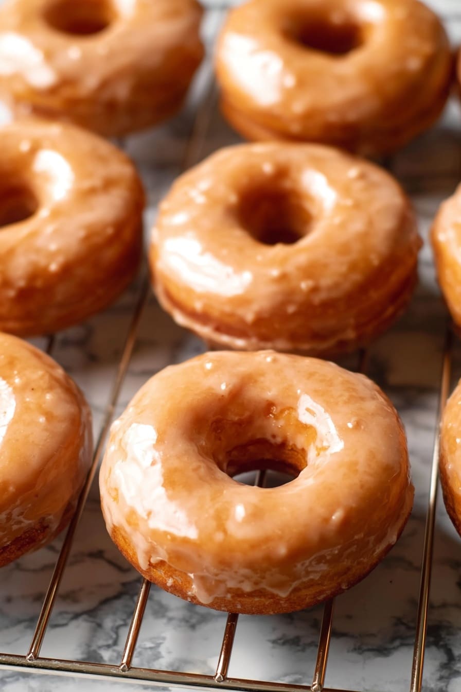 Vegan Pumpkin Donuts with Cinnamon Maple Glaze, Vegan Pumpkin Donuts, Fall Vegan Donuts, Vegan Pumpkin Dessert, Cinnamon Maple Glaze Donuts - The image shows a group of eight round donuts with a hole in the middle, all covered in a light brown glaze that looks smooth and shiny with a slightly textured surface. The donuts are placed on a metal cooling rack that has silver, straight bars running horizontally and vertically. The background features a white marbled texture. The lighting highlights the glossy glaze on the donuts, and the focus is sharp, showing the small details of the surface of the glaze and the soft texture of the donuts. photo taken with an iphone --ar 2:3 --v 7