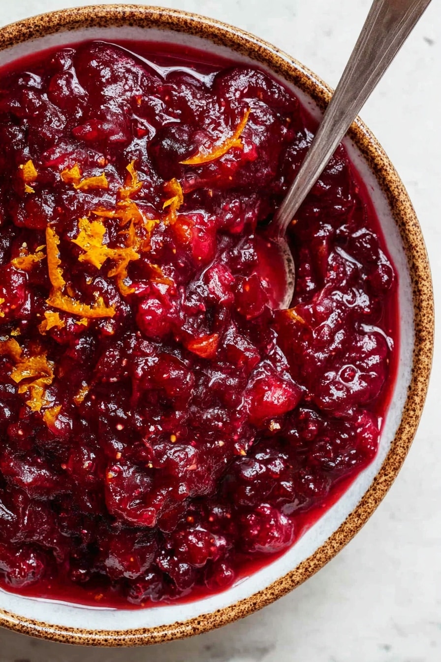 Easy Cranberry Sauce with Orange Juice, Cranberry Sauce Recipe, Holiday Cranberry Sauce, Fresh Cranberry Sauce, Orange Cranberry Sauce - A close-up view of a thick, deep red cranberry sauce with a chunky texture, showing visible pieces of fruit mixed in. Small bits of bright orange zest are scattered on top, adding color contrast. The sauce fills a white bowl with brown speckled edges, and a silver spoon is placed inside the bowl, partly submerged in the sauce. The bowl sits on a white marbled surface. photo taken with an iphone --ar 2:3 --v 7