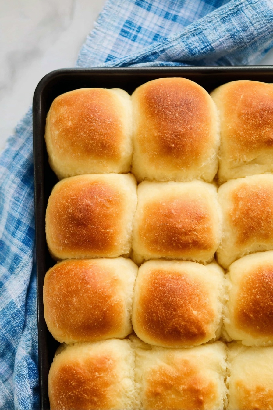 Vegan Dinner Rolls, vegan bread recipes, fluffy vegan rolls, dairy-free dinner rolls, plant-based bread - The image shows a close-up top view of sixteen golden-brown, fluffy bread rolls arranged in a 4 by 4 grid inside a black baking tray. Each roll has a slightly rounded top with a soft, smooth texture and a light crust that is golden on the upper part, fading to creamy white around the sides. The baking tray is placed on a blue and white checkered cloth on a white marbled surface. The rolls touch each other, showing their soft and puffy sides. photo taken with an iphone --ar 2:3 --v 7