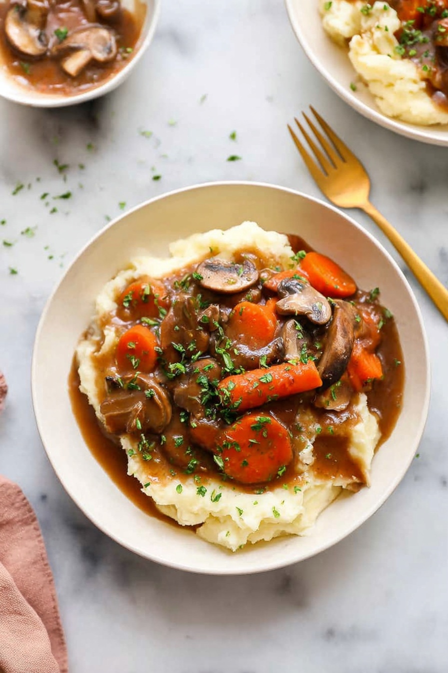 Mushroom Bourguignon, French mushroom stew, vegan mushroom stew, plant-based French dishes, mushroom stew recipe - A white bowl holds a dish with two main layers: the bottom layer is creamy mashed potatoes, soft and pale in color, covering the base of the bowl. On top is a rich brown gravy mixed with sliced mushrooms and large carrot chunks, with chopped green herbs sprinkled all over for color. A golden spoon and fork sit on the white marbled surface beside the bowl. In the background, another white bowl with the same dish is barely visible. The lighting is bright and natural, highlighting the textures and colors of the food photo taken with an iphone --ar 2:3 --v 7