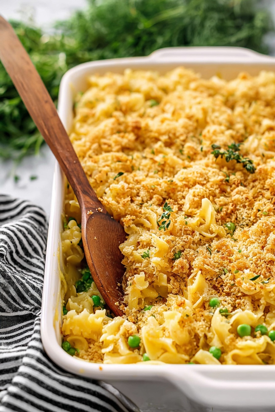 Creamy Chicken Casserole, Easy Chicken Casserole, Comforting Chicken Dinner, Cheesy Chicken Bake, Homemade Chicken Casserole - The image shows a white rectangular baking dish filled with a pasta casserole. The dish has three main layers: a bottom layer of yellow pasta, a middle layer with green peas mixed throughout, and a top layer covered with a golden brown crumbly topping that looks crispy. A wooden spoon stands inside the dish, scooping some pasta and crumbs. The background features a white marbled surface and some green herbs with a black and white striped cloth nearby. photo taken with an iphone --ar 2:3 --v 7