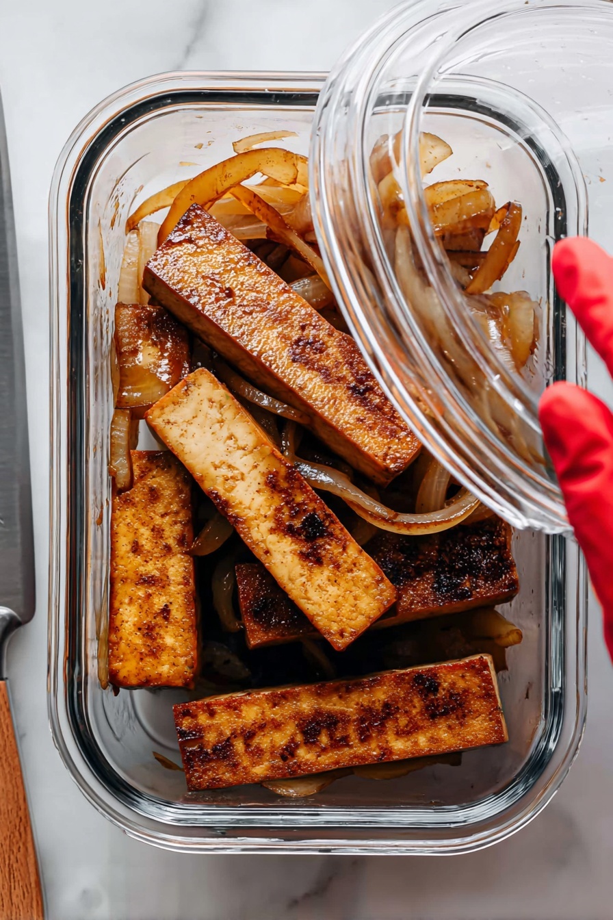 Sautéed Lime Tofu, lime tofu stir-fry, crispy tofu with lime, healthy tofu recipes, quick tofu dinner - The image shows a clear glass container holding several thick rectangular pieces of cooked tofu that are brown and have a slightly charred texture on some edges. Mixed among the tofu pieces are translucent sautéed onion slices that are light brown and curled. The container is placed on a white marbled surface, and a woman's hand with a red-gloved fingertip is lifting the clear lid. To the left, a silver knife rests on the surface partially visible. photo taken with an iphone --ar 2:3 --v 7