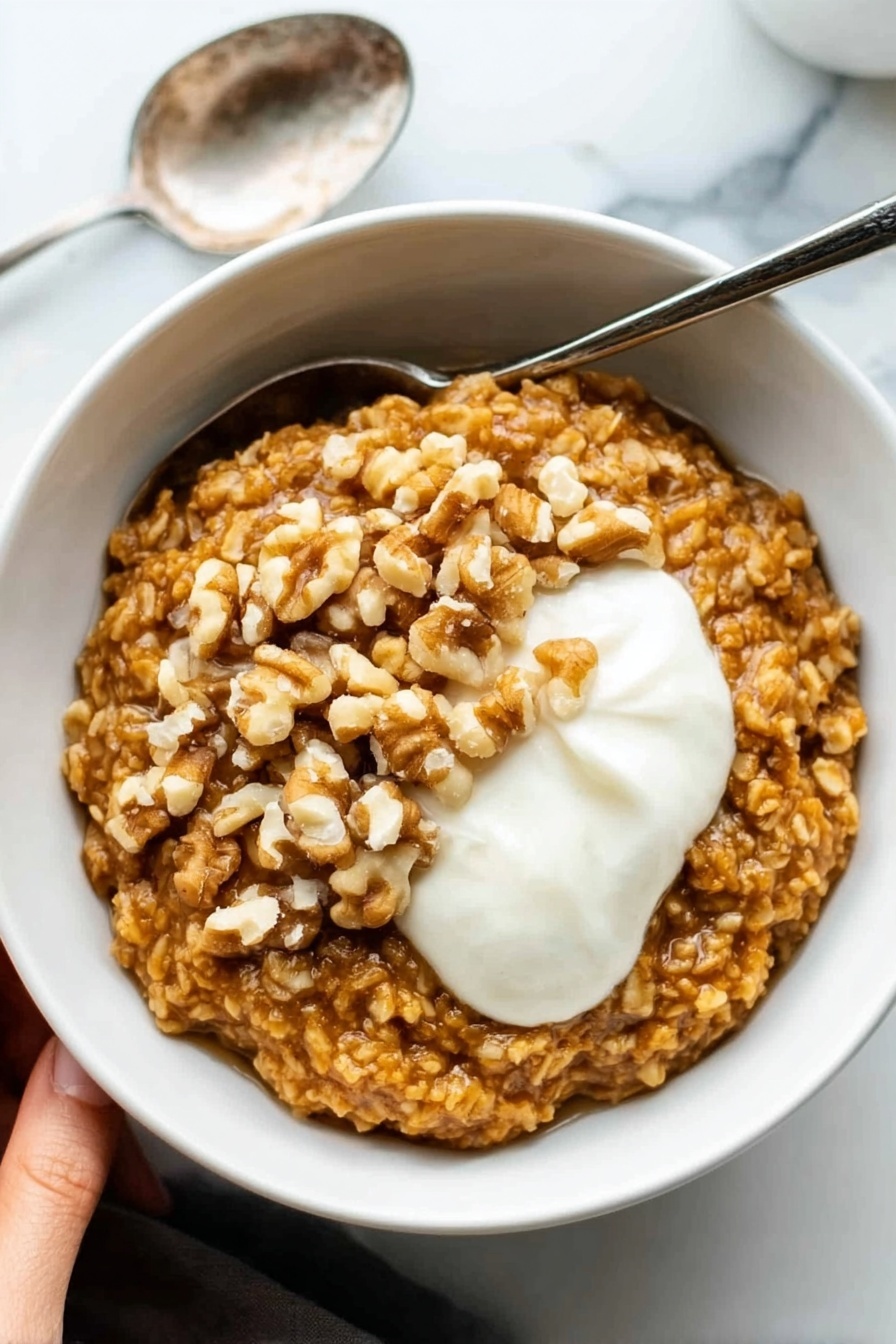 Pumpkin Oatmeal with Spiced Toppings, cozy breakfast recipes, healthy pumpkin oatmeal, seasonal fall breakfast, easy pumpkin oatmeal - A white bowl filled with thick oatmeal that is golden brown in color and has a textured look from large oats. On top of the oatmeal, there is a layer of whole walnut pieces, adding a rough, uneven surface in light brown shades. Next to the walnuts, there is a dollop of smooth, white cream sitting on the right side of the bowl. The bowl is set on a white marbled surface, and a silver spoon rests next to it on the left. In the upper left corner, a woman's hand is visible holding the bowl. Photo taken with an iphone --ar 2:3 --v 7