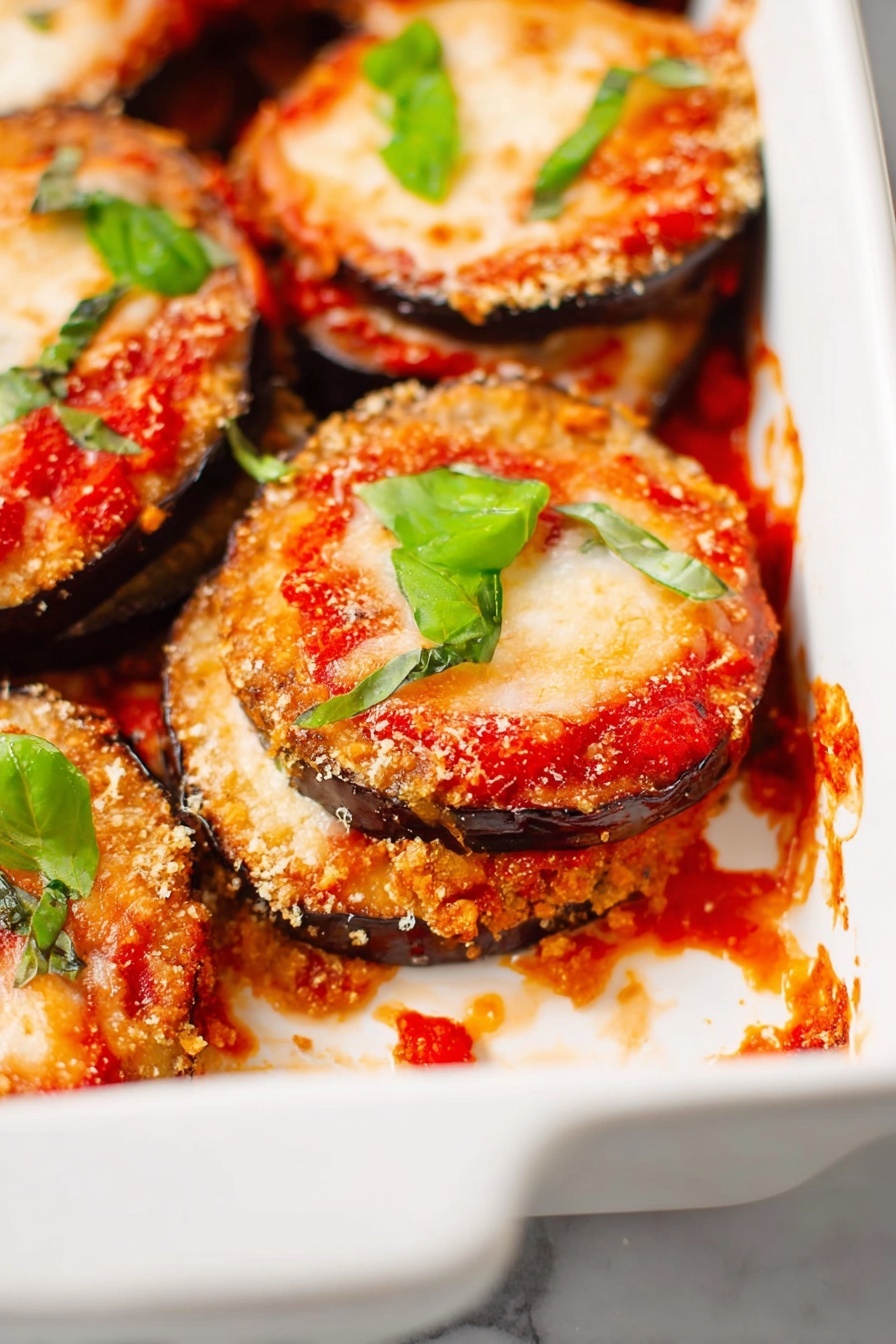 Vegan Eggplant Parmesan, Plant-Based Eggplant Parmesan, Vegan Italian Dinner, Healthy Eggplant Parmesan, Vegan Cheese Eggplant Dish - The image shows a white ceramic baking dish filled with several layered stacks of eggplant Parmesan. Each stack has a bottom layer of crispy, golden-brown eggplant slices with a rough texture, topped with rich red tomato sauce, then a layer of melted pale orange cheese that is slightly bubbly and browned in spots. Bright green basil leaves are scattered on top of the stacks and around the dish for color contrast. There are some tomato sauce spills and melted cheese crumbs inside the dish on a white marbled surface. Photo taken with an iphone --ar 2:3 --v 7