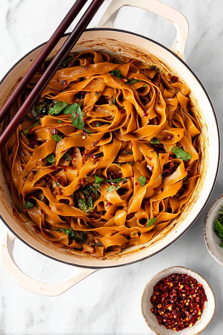 Spicy Chili Garlic Noodles, chili garlic noodle dish, spicy Asian noodles, quick garlic noodle recipe, fiery stir-fry noodles - A white bowl filled with wide flat noodles coated in a shiny reddish-brown sauce is shown from above. The noodles are mixed and curled inside the bowl with visible pieces of fresh green onions and chopped cilantro scattered on top. There are small white sesame seeds and dark chili flakes sprinkled over the noodles. A pair of dark wooden chopsticks rests on the edge of the bowl leaning inward. In the blurred background, two small white bowls hold red chili flakes and more chopped green onions. The surface is a white marbled texture. photo taken with an iphone --ar 2:3 --v 7