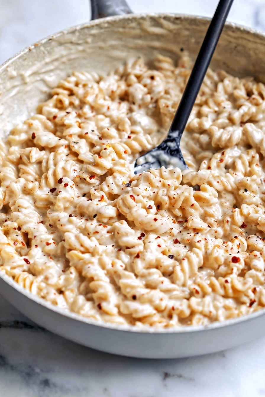 Creamy Cottage Cheese Pasta Sauce, healthy pasta sauces, protein-packed pasta recipes, easy pasta sauce, homemade pasta sauce with cottage cheese - The image shows a large gray bowl filled with a creamy, light beige pasta dish. The pasta is rotini, with a spiral shape, fully coated with a thick, smooth sauce that has a cheesy and slightly speckled texture. A woman's hand holding a metal spoon is stirring the pasta, lifting a spoonful from the center. The bowl sits on a surface with a white marbled texture. The overall look is warm and creamy, with the pasta fully covered in the sauce to give it a rich appearance. photo taken with an iphone --ar 2:3 --v 7