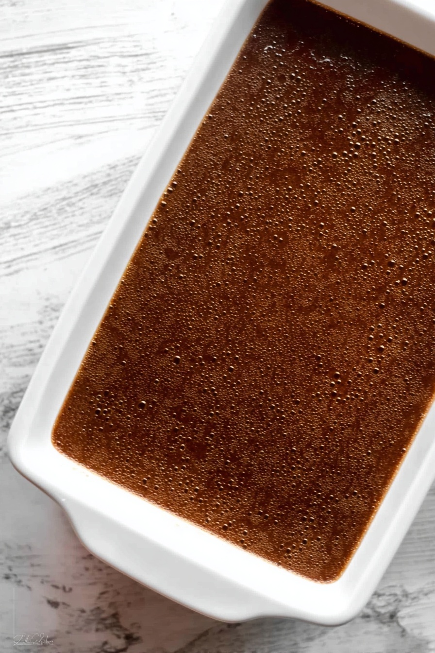 Gingerbread Cake with Butterscotch Sauce, gingerbread cake recipe, spiced gingerbread cake, cozy holiday dessert, easy gingerbread cake - The image shows a white rectangular baking dish filled with a smooth, dark brown dessert that has a few small bubbles on the surface, giving it a slightly textured look. The dish sits on a white marbled surface with visible veins. The dessert has a shiny, even top layer that reflects some light, indicating a moist texture beneath. Photo taken with an iphone --ar 2:3 --v 7