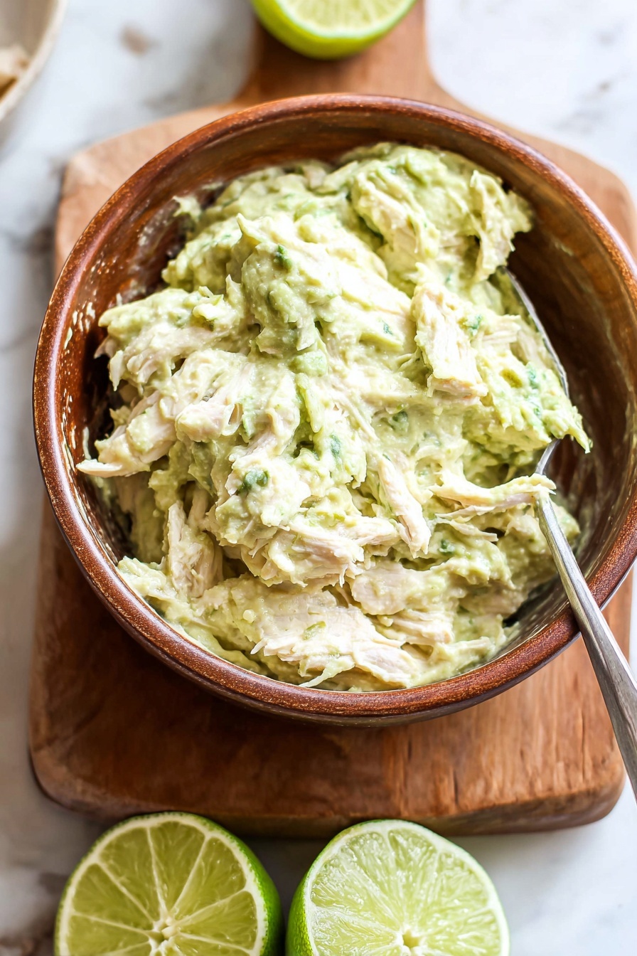 Healthy Greek Yogurt Avocado Chicken Salad, healthy chicken salad, avocado chicken salad, Greek yogurt chicken salad, light healthy lunch ideas - A brown bowl filled with a creamy, light green mixture that has a chunky texture with visible pieces of shredded white meat mixed inside. The bowl is placed on a wooden board, which rests on a white marbled surface. Behind the bowl, there are two halves of a lime, one facing up showing the green juicy interior, and the other turned to the side. A metal spoon is partially inserted into the mixture in the bowl. The scene is bright and fresh. photo taken with an iphone --ar 2:3 --v 7