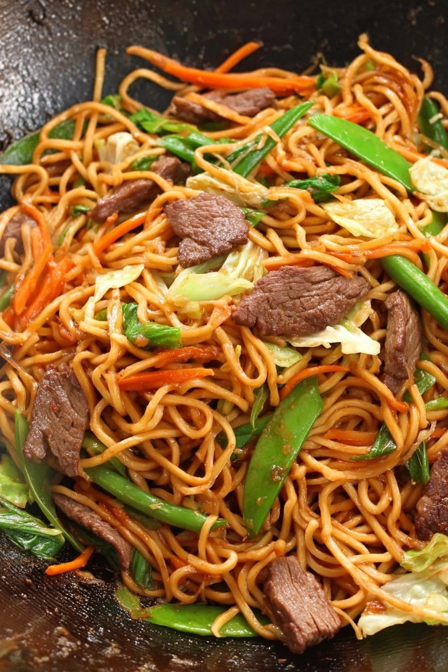 Easy Beef Lo Mein, beef noodle stir-fry, quick Asian dinner, savory beef recipes, family-friendly Chinese dishes - This image shows a close-up of a wok filled with stir-fried noodles, meat slices, and mixed vegetables. The noodles are long and orange-brown, forming the main layer twisted together. Scattered throughout are medium-brown pieces of cooked meat, adding a rich texture. Bright green vegetables like sliced green onion and snap peas are placed evenly, with thin strips of orange carrot and light green cabbage mixed in, adding new colors and crunch. The surface inside the wok is dark and slightly shiny, highlighting the fresh, vibrant stir-fry. photo taken with an iphone --ar 2:3 --v 7
