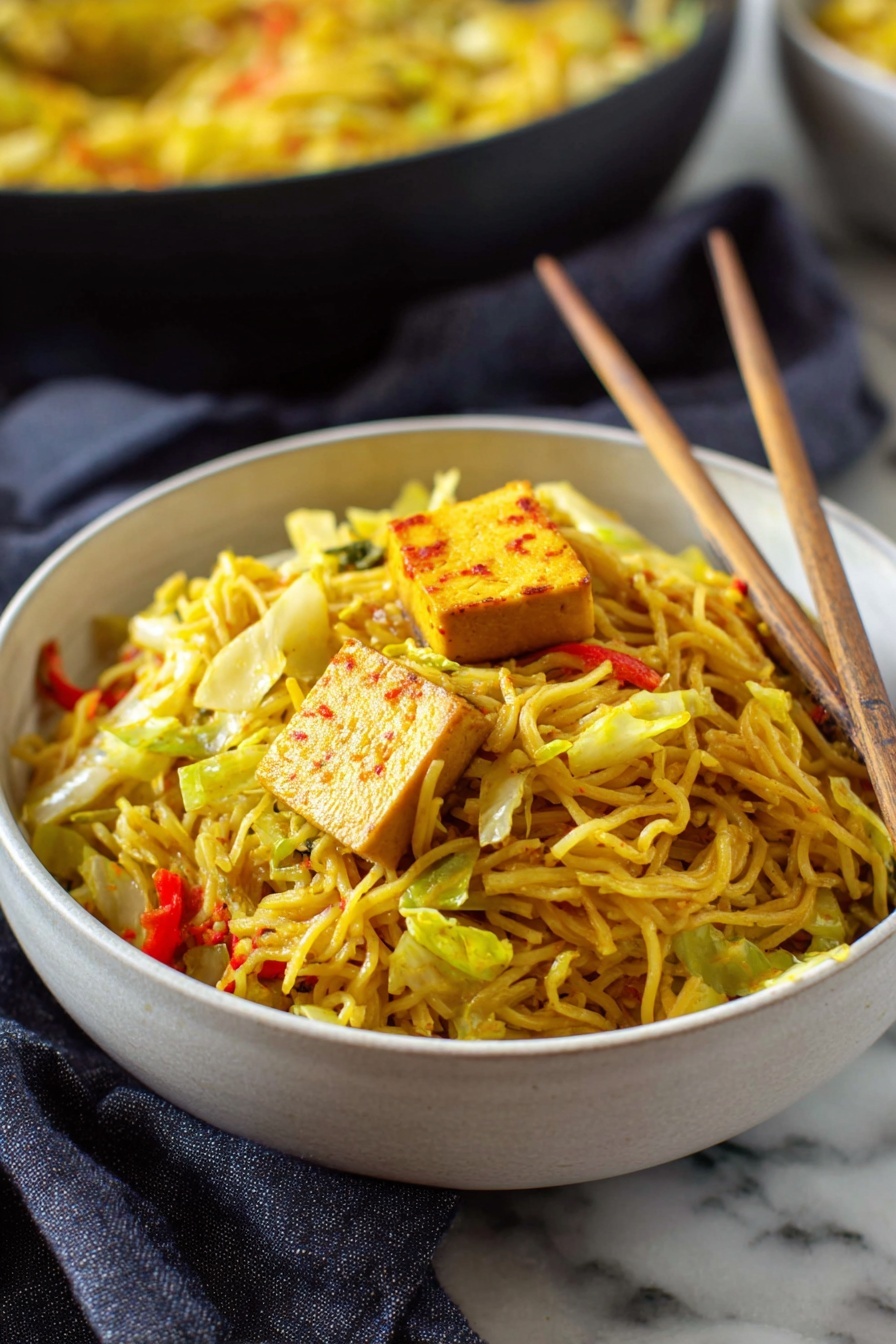 Easy Singapore Noodles with Curry and Vegetables, Singapore Noodles recipe, quick Asian noodle dish, flavorful vegetable stir-fry, easy dinner ideas with rice noodles - A deep white bowl full of thin yellow noodles mixed with pieces of green cabbage and small bits of red bell pepper, topped with a square piece of light brown tofu with reddish edges. Light wooden chopsticks rest inside the bowl, leaning against the noodles on the right side. The bowl sits on a white marbled surface with a dark gray cloth nearby. In the background, there is another bowl filled with more noodles, blurred slightly. Photo taken with an iphone --ar 2:3 --v 7