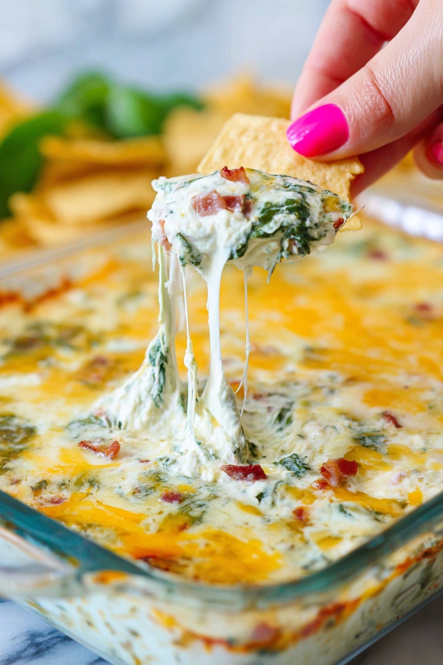 Bacon Spinach Artichoke Dip, cheesy bacon spinach artichoke appetizer, creamy bacon spinach dip, savory bacon artichoke dip, easy party dip recipes - A white square glass dish shows a baked dip with a top layer of melted golden-yellow cheese, lightly browned in spots. Under the cheese, thick creamy white sauce mixed with green spinach leaves and small bits of reddish-brown bacon is visible. A woman's hand with pink painted nails holds a triangular beige chip pulling up a string of stretchy melted cheese from the dish, showing the layers clearly. The background is blurry with hints of green and more chips. The whole scene is set on a white marbled surface. photo taken with an iphone --ar 2:3 --v 7