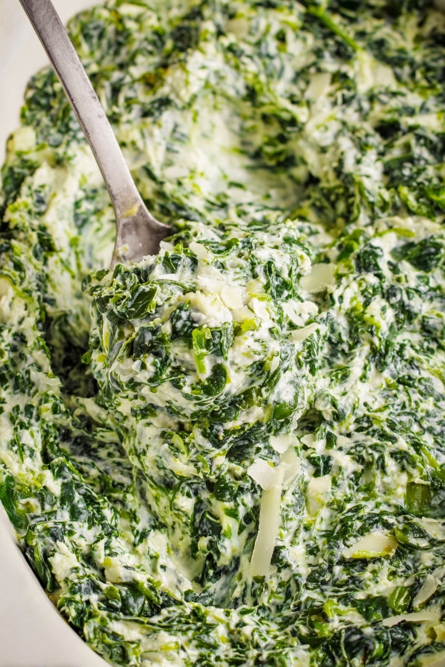 Creamed Spinach, Creamed Spinach Side Dish, Easy Creamed Spinach, Gourmet Spinach Recipes, Creamed Spinach with Cheese - A close-up view of creamy spinach dip with visible layers of finely chopped green spinach mixed with white creamy sauce and small pieces of onion. The texture looks thick and rich with bits of grated white cheese sprinkled on top. A metal spoon scoops up a generous portion of the dip, showing the mix's dense, slightly chunky consistency. The dish sits on a white marbled surface that contrasts with the deep green and white colors of the dip. Photo taken with an iphone --ar 2:3 --v 7