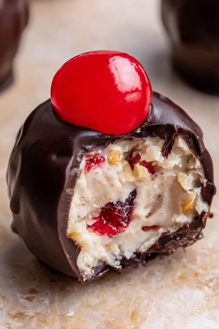 Cherry Bourbon Balls, Bourbon Balls with Cherries, No-Bake Boozy Treats, Holiday Chocolate Bites, Easy Alcohol Infused Snacks - The image shows a close-up view of a small dessert ball with three layers. The outside layer is dark, smooth, and shiny chocolate coating covering the entire ball. Inside, the second layer is light-colored, creamy, and soft with some small red and golden brown bits mixed inside, giving it a textured look. On top of the chocolate layer, there is a bright red, glossy half of a cherry placed in the center. The ball sits on a light brown surface with a white marbled texture softly blurred in the background. photo taken with an iphone --ar 2:3 --v 7