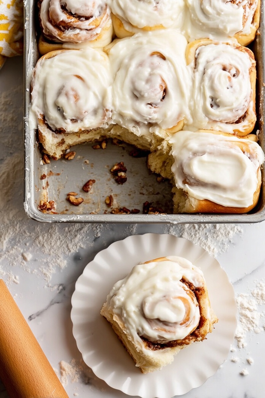 Soft Apple Cinnamon Rolls, apple cinnamon rolls recipe, easy apple cinnamon rolls, warm cinnamon apple rolls, flavorful breakfast rolls - A metal baking dish holds six soft cinnamon rolls, with one removed showing the bottom layer coated in a sticky cinnamon filling and bits of nuts. Each roll has a creamy white icing thickly spread on top, swirled smoothly over the soft golden brown dough, with visible cinnamon spirals peeking through under the icing. One cinnamon roll sits on a small white scalloped plate in the lower right corner, showing its fluffy layered inside. The background is a white marbled surface lightly dusted with flour, with a wooden rolling pin nearby. photo taken with an iphone --ar 2:3 --v 7
