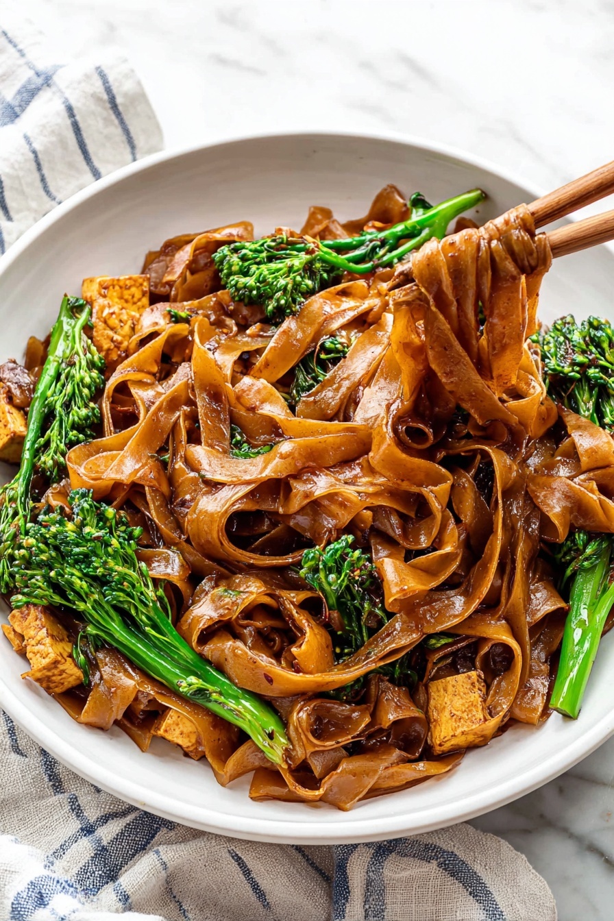 Quick Thai Pad See Ew Stir Fry, Thai noodle stir fry, authentic Pad See Ew, easy Thai stir fry recipe, quick Asian noodle dish - A white bowl is filled with wide, flat noodles coated in a shiny dark brown sauce, layered thick, with some noodles lifted by wooden chopsticks to the right side of the image. Mixed in are pieces of light brown cooked tofu and bright green broccolini with textured small florets. The bowl sits on a white cloth with blue stripes on a white marbled surface. The colors show a contrast between the dark noodles, green vegetables, and pale tofu, creating a rich and fresh look. photo taken with an iphone --ar 2:3 --v 7