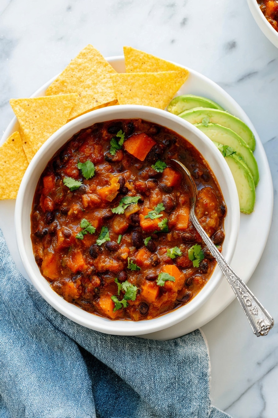 Vegetarian Chili, Best Vegetarian Chili, Hearty Vegetarian Chili, Easy Vegetarian Chili Recipe, Healthy Vegetarian Chili - A white bowl filled with thick chili containing black beans, orange carrot pieces, and chunks of sweet potato in a reddish-brown sauce, garnished with green cilantro leaves. On the right side inside the bowl are thin slices of light green avocado sprinkled with cilantro. The bowl sits on a white plate arranged with five triangular yellow tortilla chips around the left side. A silver spoon with a decorative handle rests inside the chili. The setting has a white marbled texture surface with a slightly crumpled blue cloth napkin next to the bowl. Photo taken with an iphone --ar 2:3 --v 7