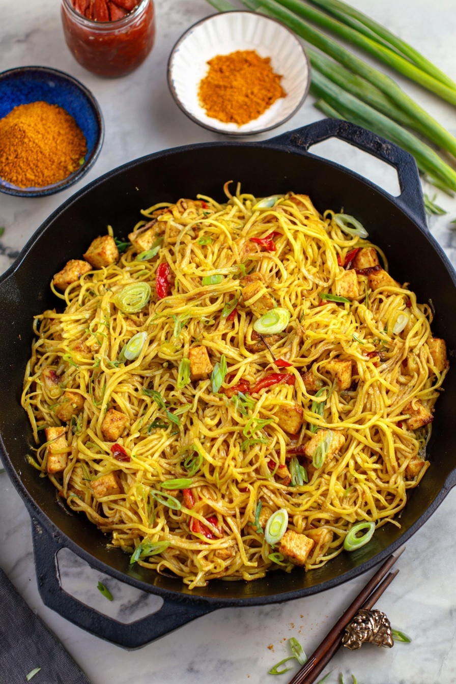 Easy Singapore Noodles with Curry and Vegetables, Singapore Noodles recipe, quick Asian noodle dish, flavorful vegetable stir-fry, easy dinner ideas with rice noodles - A black cast iron pan filled with several layers of yellow noodles mixed with small pieces of grilled tofu that show a light brown char, scattered with thin slices of green onions and small bits of red bell pepper. The noodles have a slight shine and look seasoned. Around the pan, there is a white bowl with orange powder, a jar with red sauce, a blue bowl with chopsticks placed on the side, and some green onion stalks on a white marbled surface. The scene has a mix of greens and yellows that make the dish look fresh and tasty photo taken with an iphone --ar 2:3 --v 7