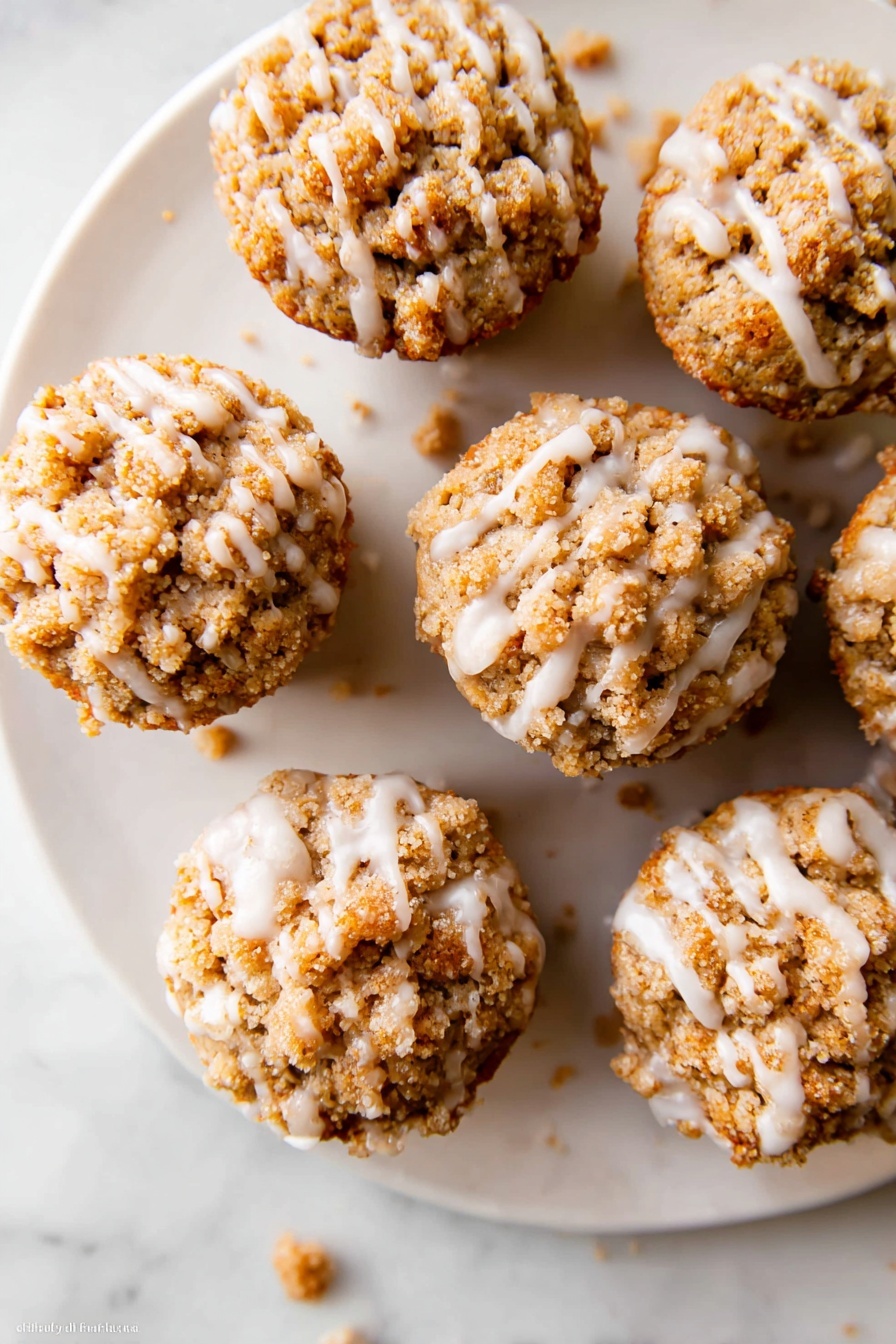 Pumpkin Crumb Muffins with Maple Icing, pumpkin muffin recipes, autumn pumpkin muffins, maple icing bakery treats, cozy fall muffin ideas - The image shows a white plate with six small muffins arranged in a loose circle on a white marbled surface. Each muffin has a crumbly, light brown streusel topping that covers the whole top and looks crunchy. A thin layer of white icing is drizzled unevenly over the muffins, creating shiny lines that drip slightly onto the plate. A few crumbs are scattered around the plate and on the marbled surface. The muffins look moist and textured with a mix of crumbly and smooth parts. Photo taken with an iphone --ar 2:3 --v 7