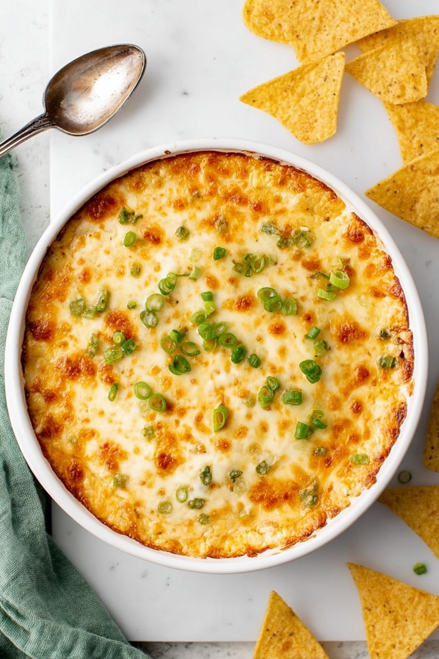 Crab Rangoon Dip, Crab Rangoon Dip with Crispy Wontons, cheesy crab dip, savory appetizer dip, crab and cream cheese dip - A round white baking dish filled with a golden, bubbly cheese layer on top, showing patches of light brown where the cheese is slightly toasted, scattered with small rings of bright green chopped scallions. The entire dish sits on a white marbled surface with a vintage silver spoon to the side, broken and whole triangular yellow tortilla chips nearby, and a soft green cloth partly visible at the edge. Photo taken with an iphone --ar 2:3 --v 7